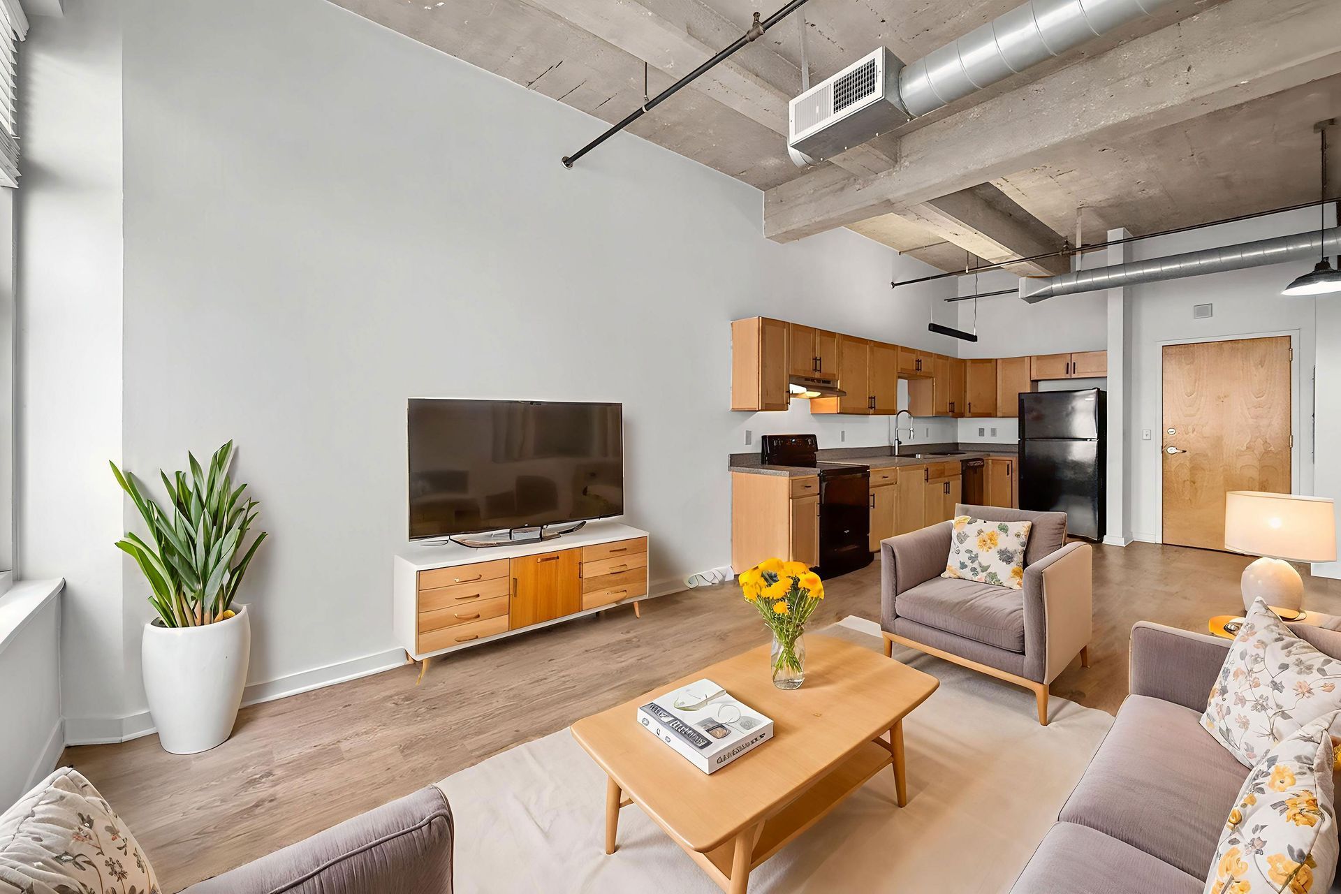 A modern open-concept apartment with a living room, light wood furniture, grey armchairs, a TV, and a kitchen area.