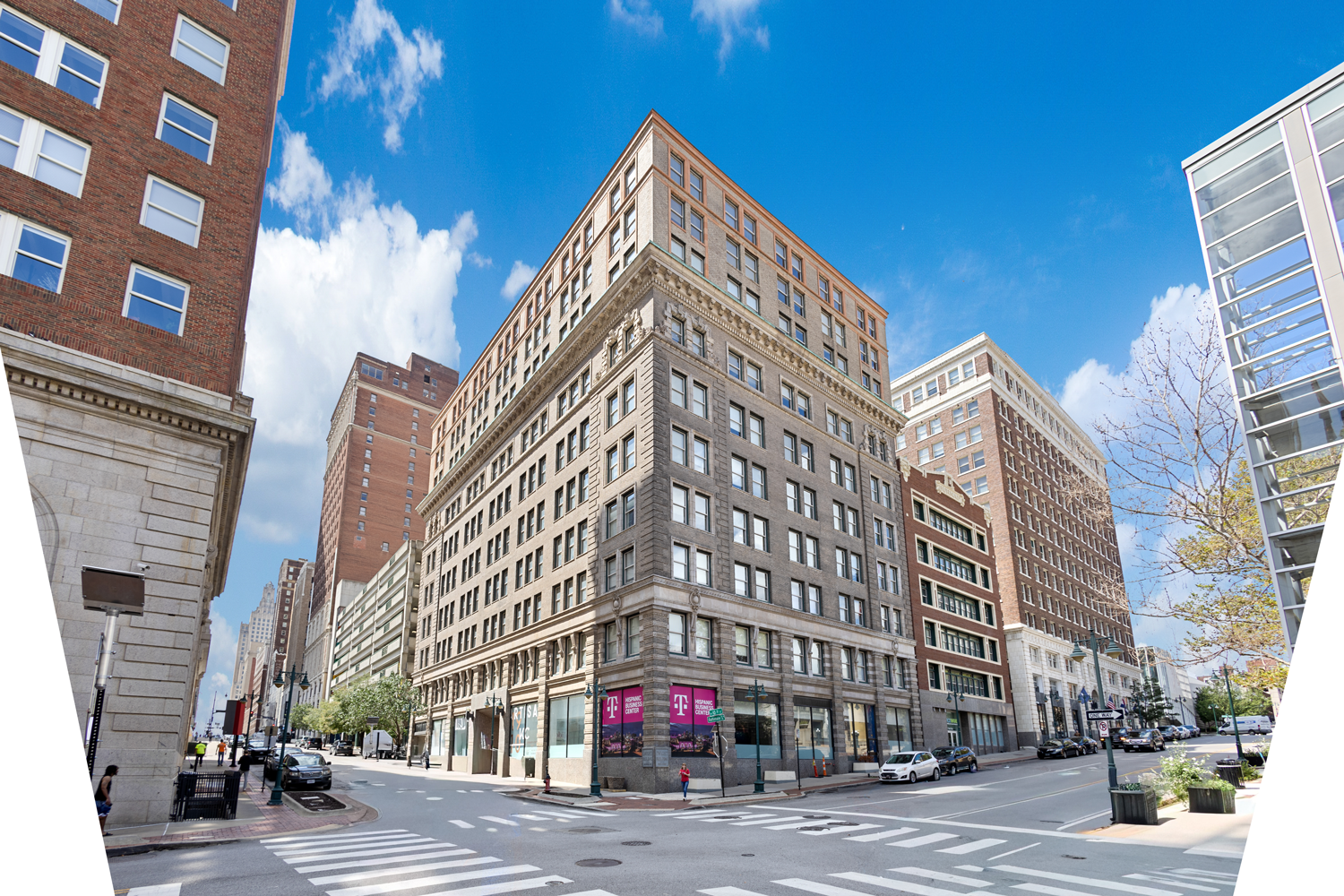 A low-angle view of a multi-story, grey stone commercial building on a street corner under a bright blue sky.