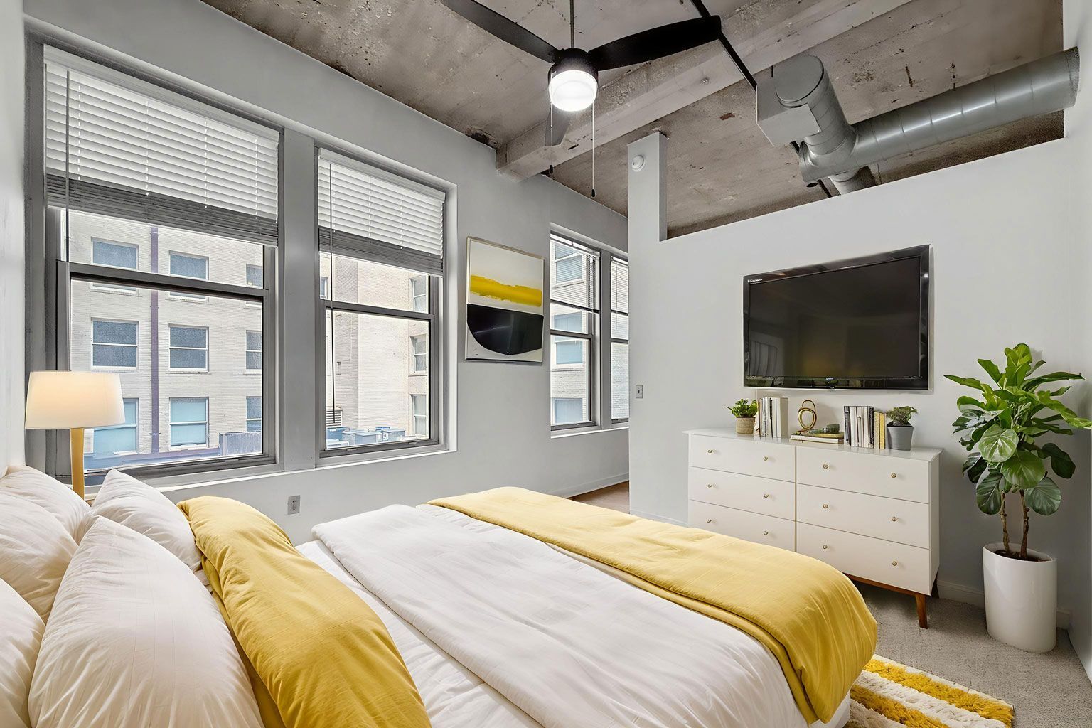 A modern bedroom with a bed featuring yellow bedding, a dresser with a TV above it, a potted plant, and exposed ceiling.
