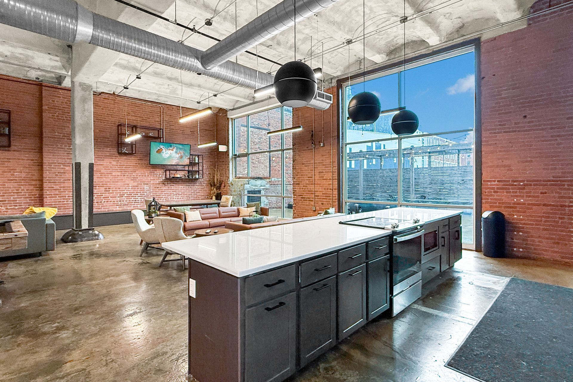 An open-concept loft with a large kitchen island, industrial brick walls, exposed ductwork, and floor-to-ceiling windows.