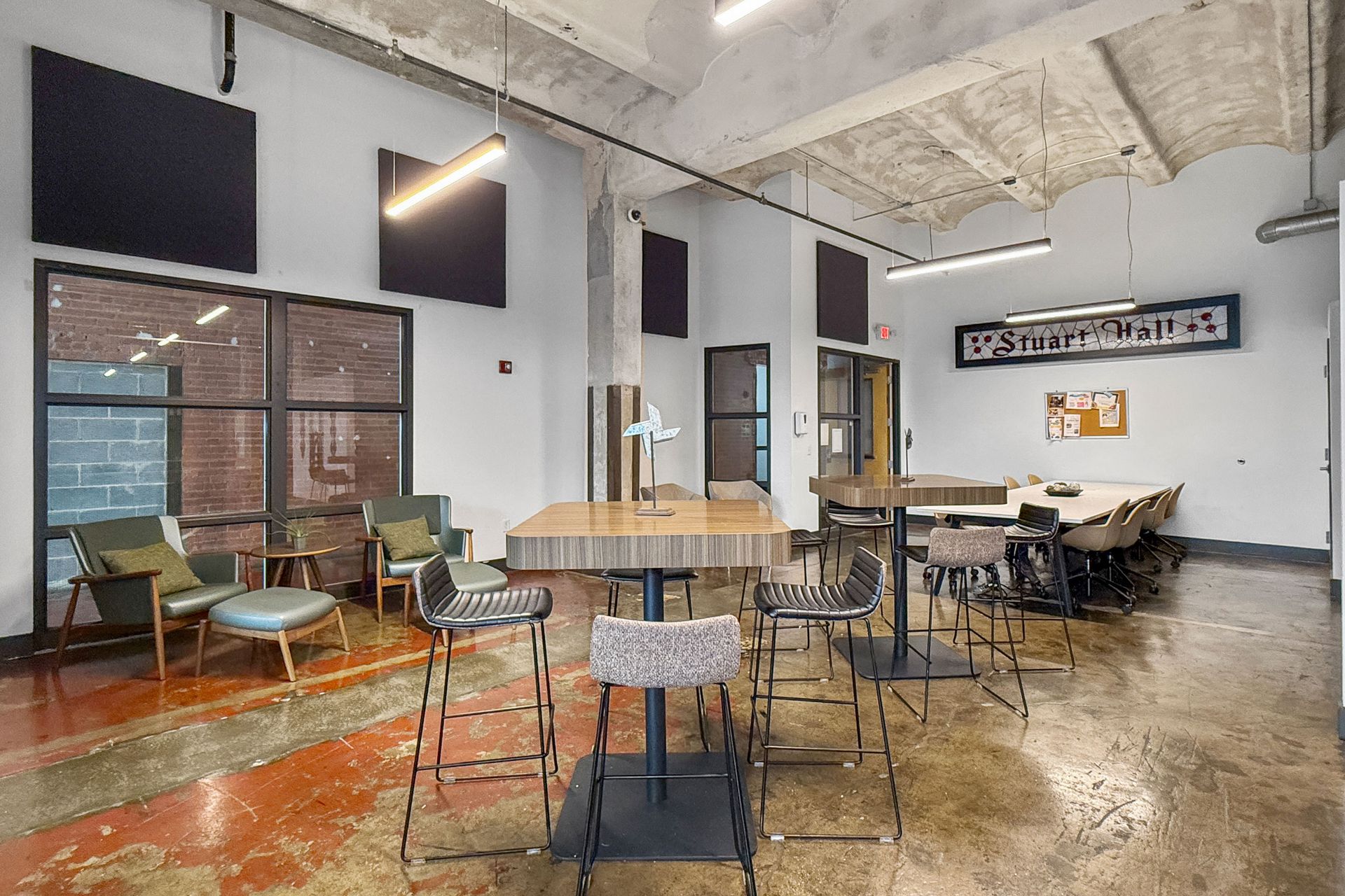 Modern industrial office lobby with a lounge area, tall pub tables, and a long conference table on a stained concrete floor.