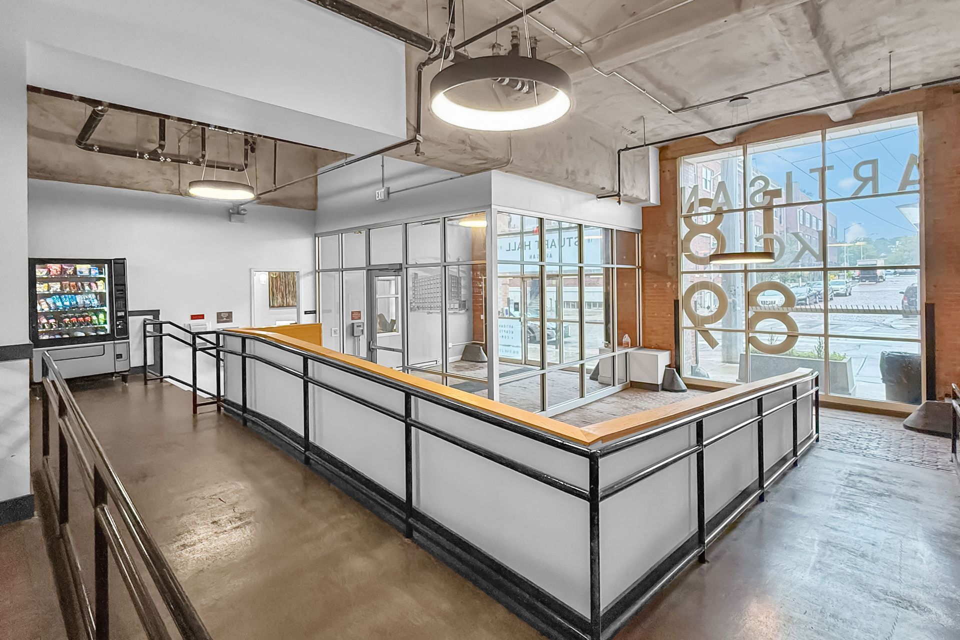 An industrial-style loft office interior with a glass-enclosed meeting room, a snack vending machine, and large windows.