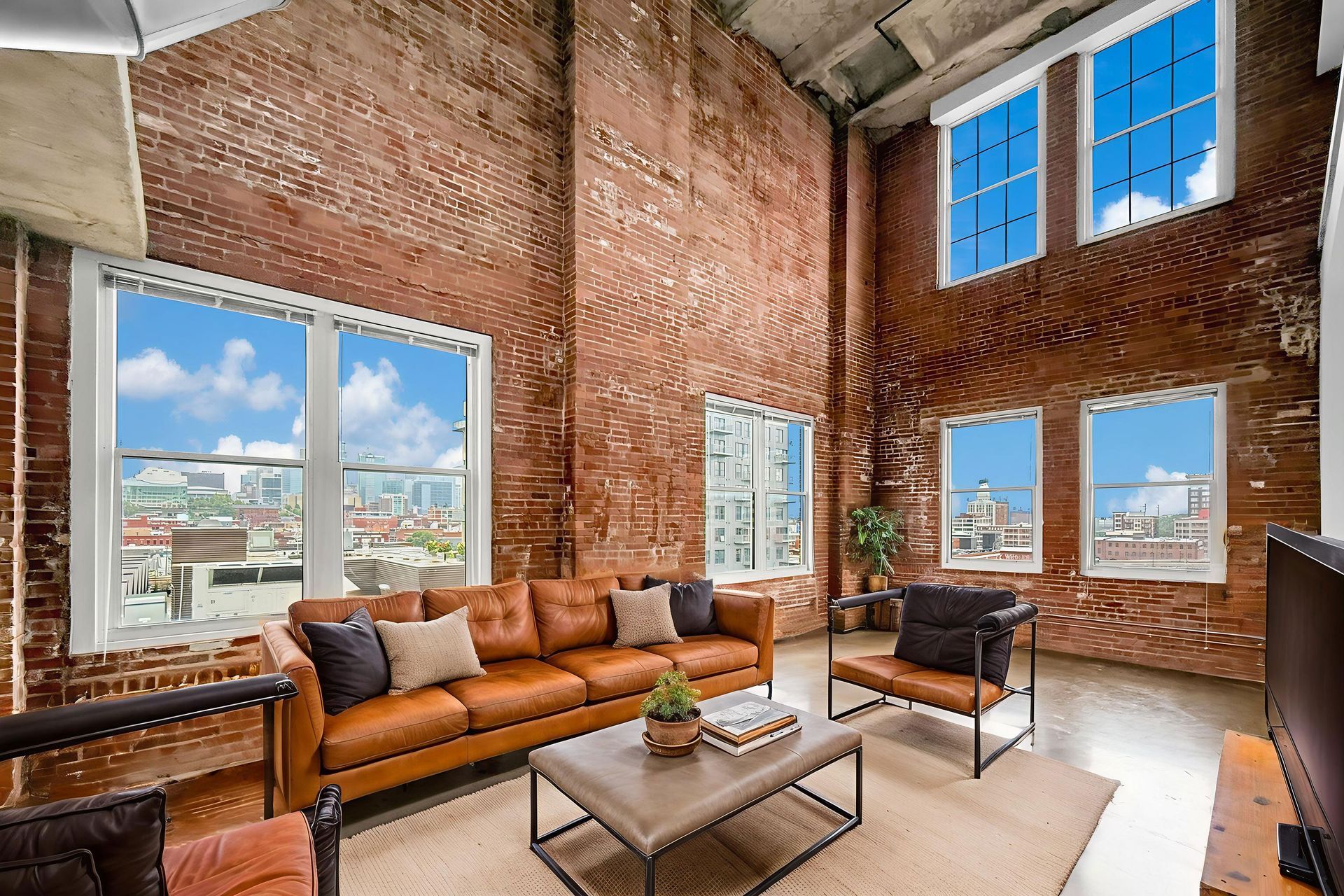 A sunlit industrial loft with exposed brick walls, high ceilings, a leather sofa, coffee table, and large windows.
