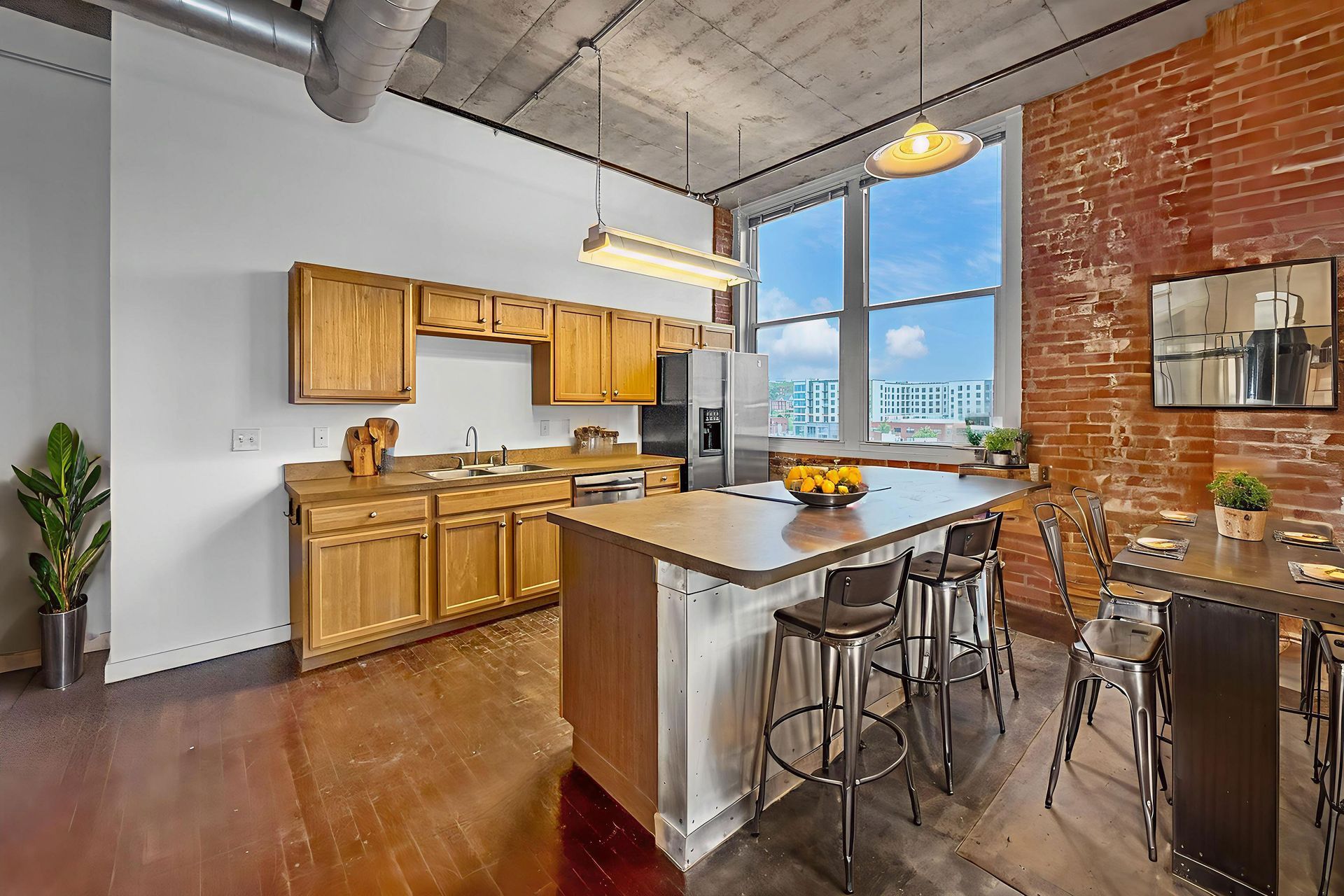 A modern kitchen with oak cabinets, a central island with bar stools, an exposed brick wall, and large windows.