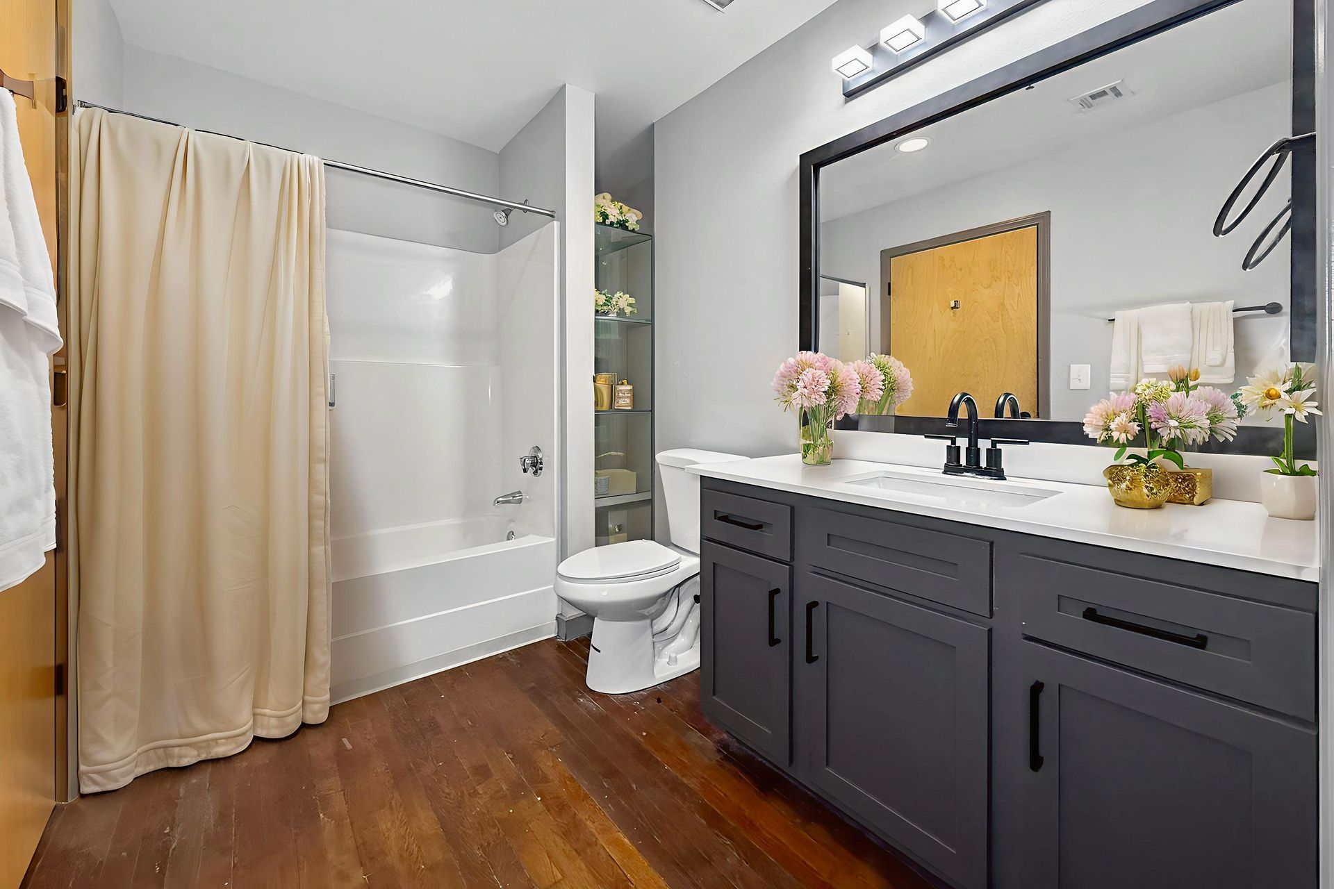 A bathroom with dark grey vanity, white countertops, a walk-in tub with a beige curtain, and wood-look flooring.