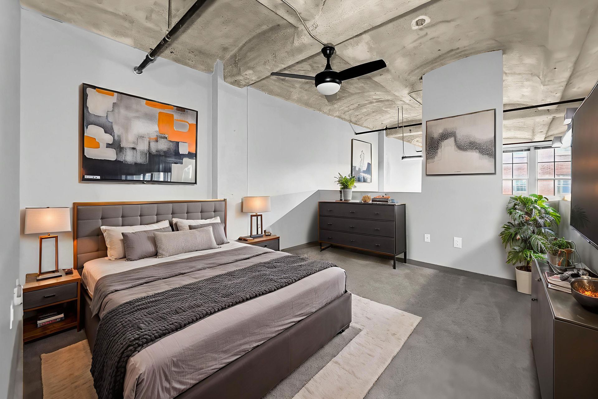 A bedroom with a grey tufted bed, concrete ceiling, black fan, dresser, and abstract art in a modern industrial apartment.