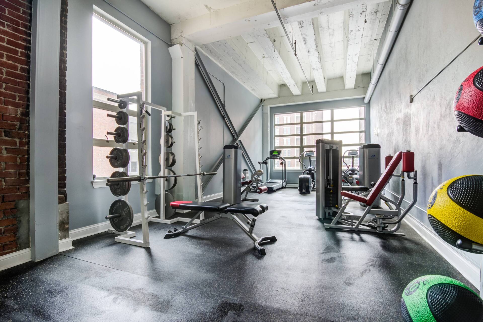 An indoor gym with workout equipment, including a weight rack, weight bench, and medicine balls, against a brick wall.
