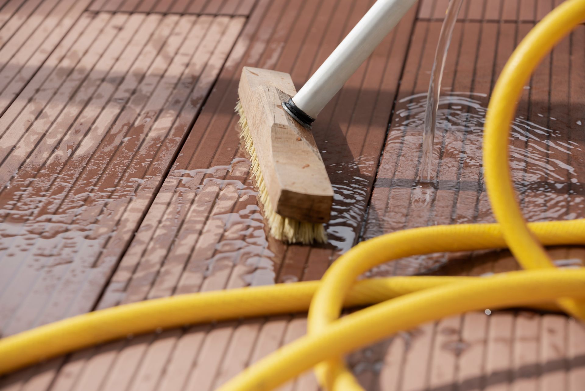 A broom is being used to clean a wooden floor next to a yellow hose