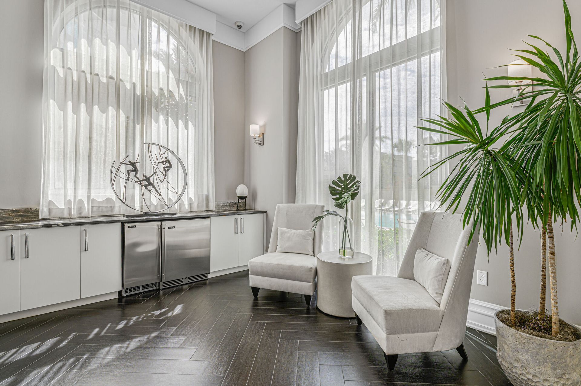Bright modern living area with tall windows, sheer curtains, two white chairs, a round table, and a large potted plant.