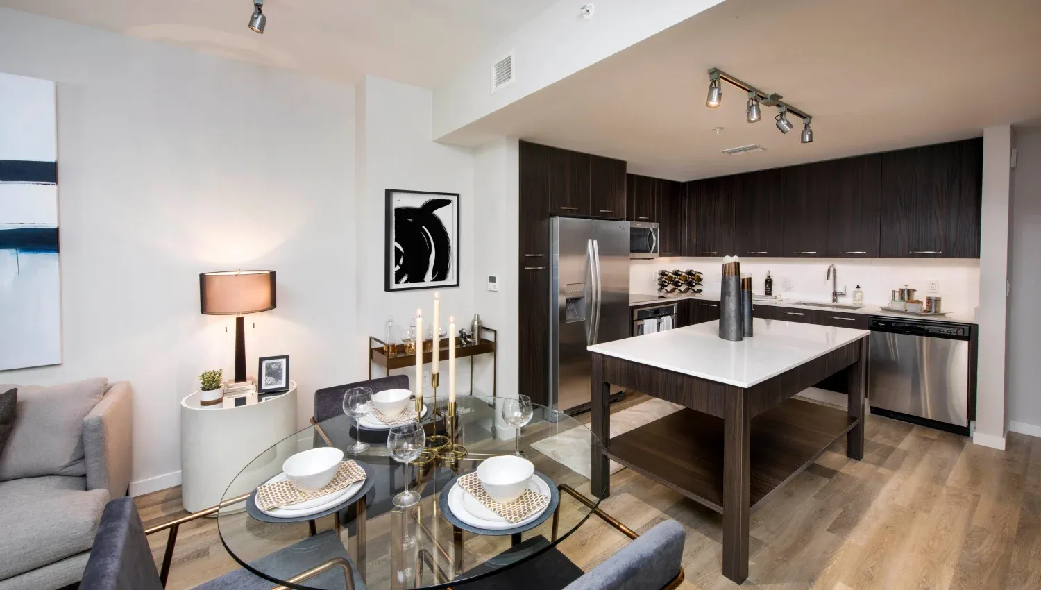 Open-concept kitchen with dark wood cabinets, stainless steel appliances, and a glass dining table in a modern apartment.