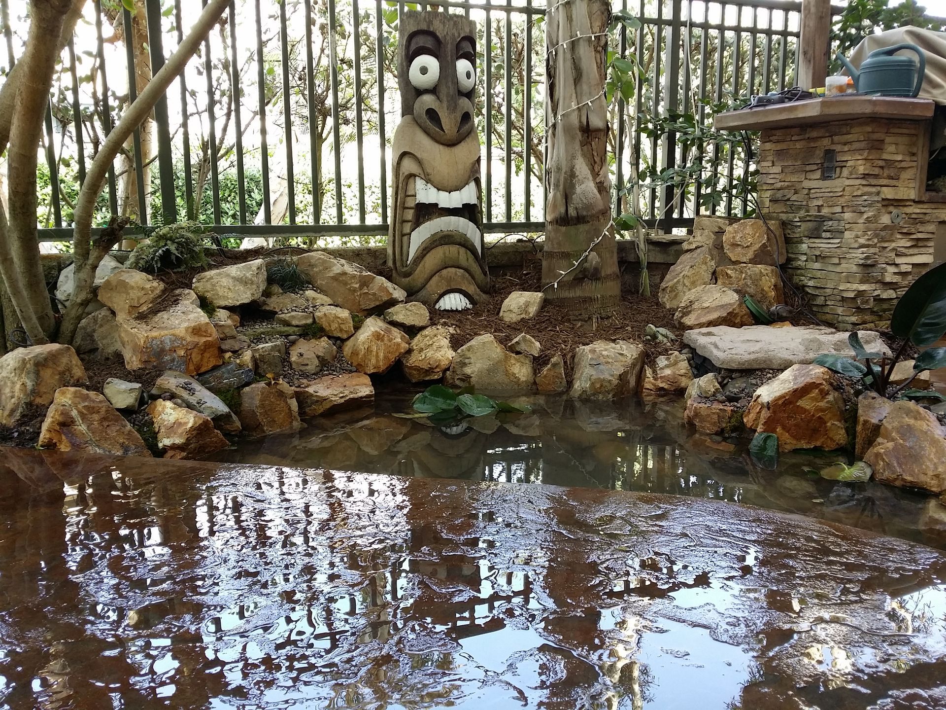 Tiki statue beside a small pond surrounded by rocks in a garden. Reflection visible in the water.