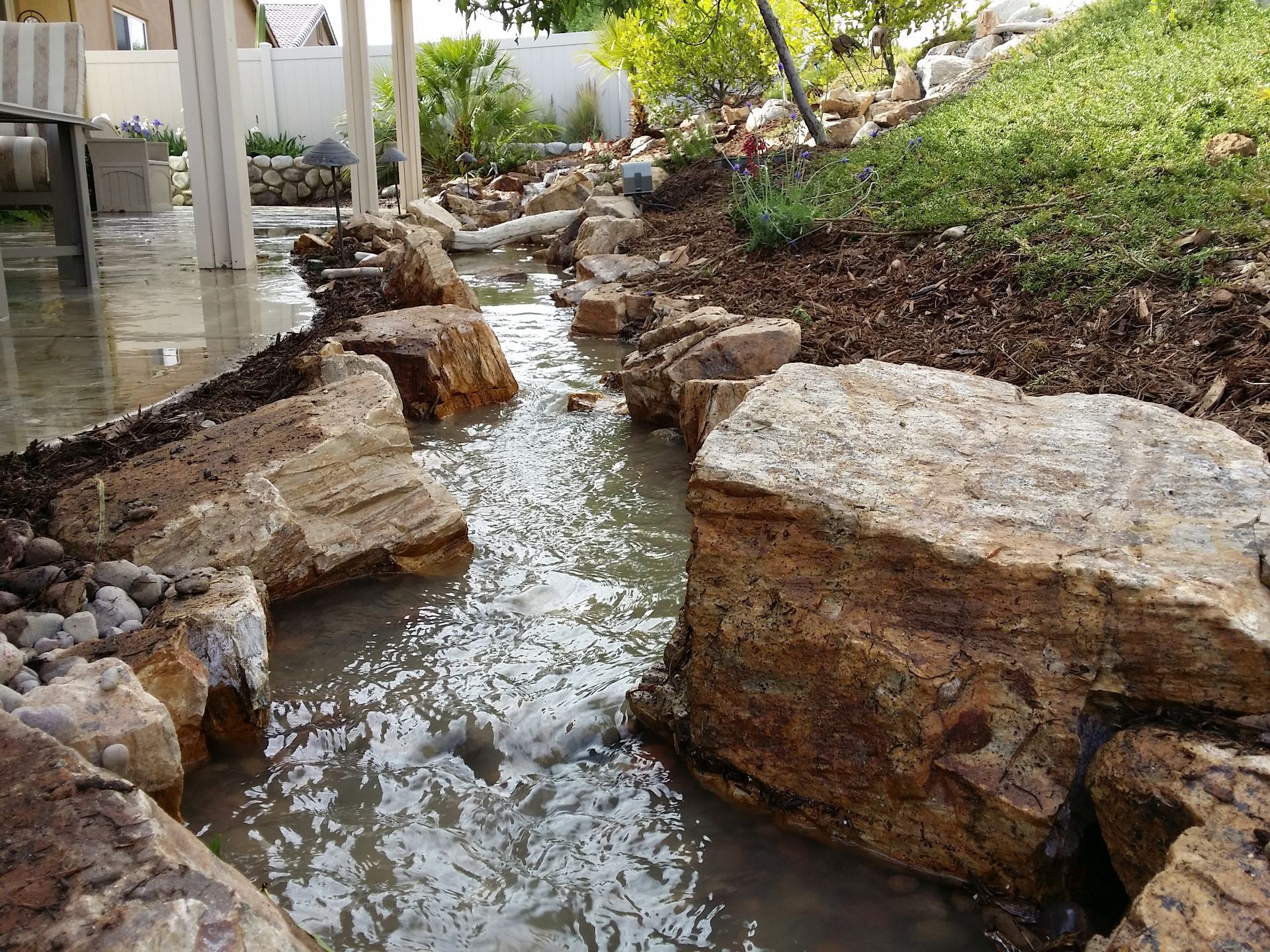 Water flows through a rock-lined stream in a landscaped yard.
