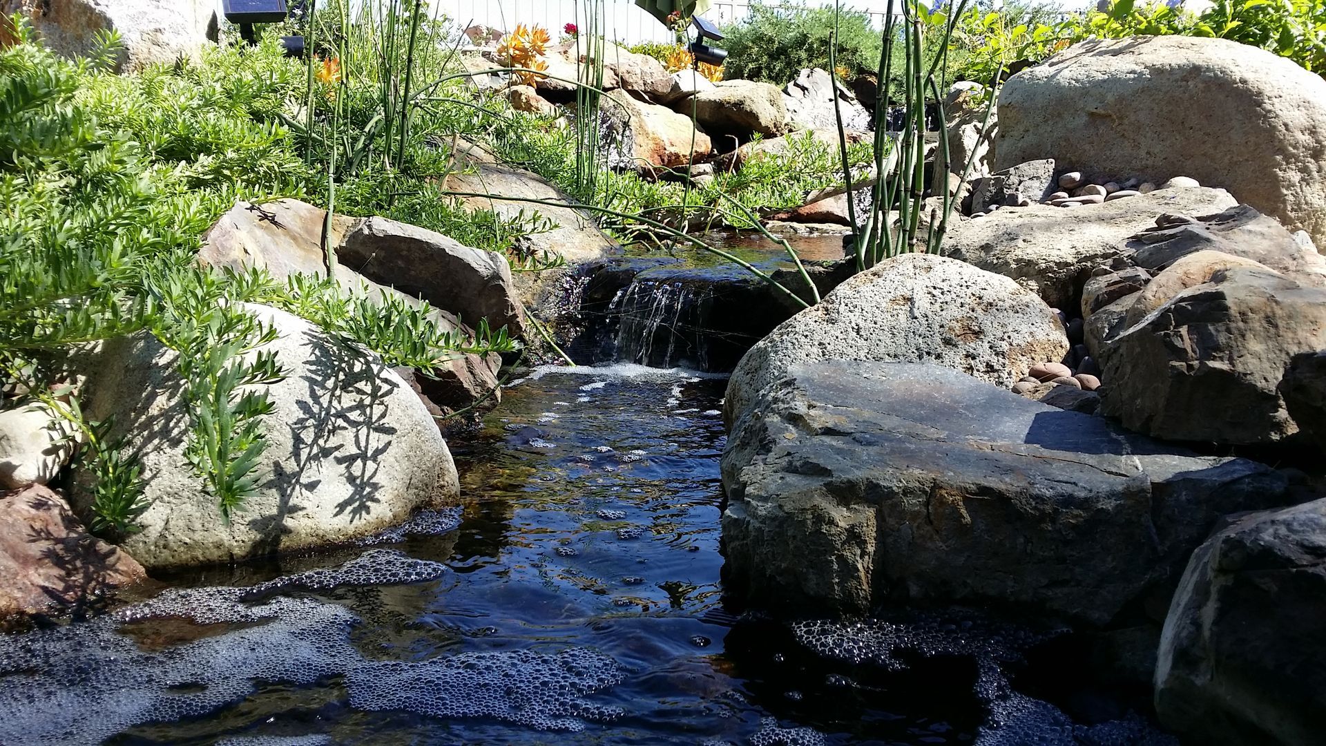 Small waterfall flowing through rocks, surrounded by green plants in a garden setting.