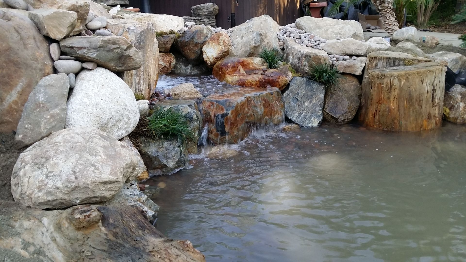 Waterfall cascading into a small pond surrounded by rocks and a wooden stump.