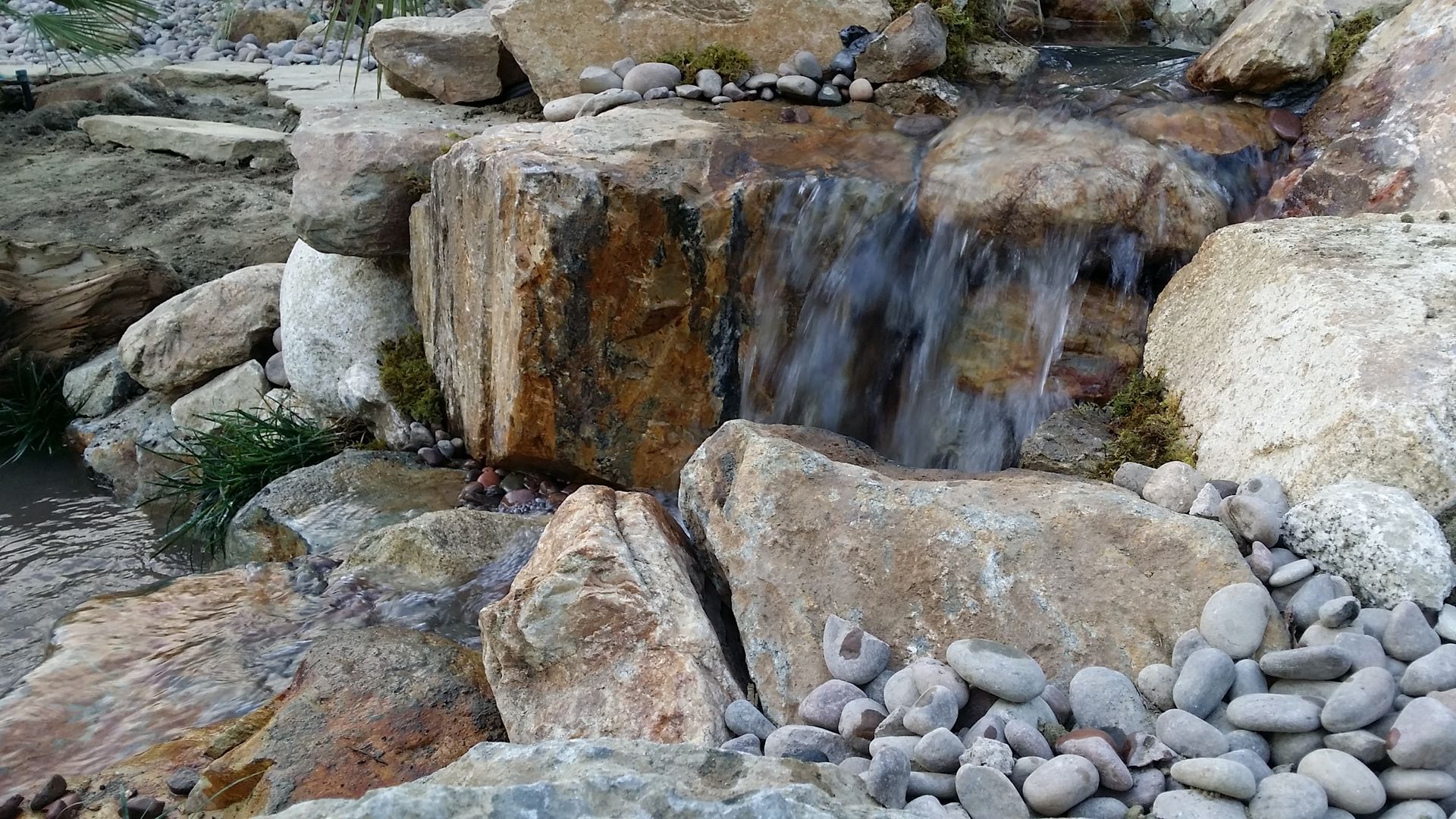 Water cascades over large, tan-colored rocks, forming a small waterfall in a garden setting.
