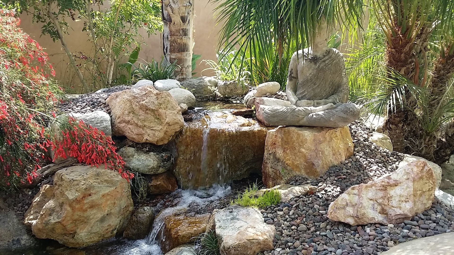 A small, rocky waterfall in a garden, with a seated Buddha statue on a rock.