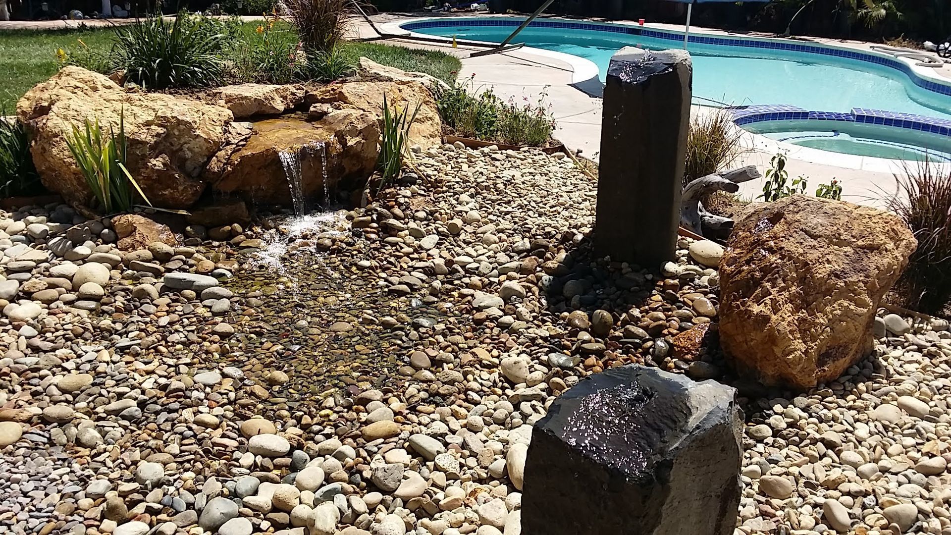 Water feature with small waterfall and stone fountain in a backyard with a pool.