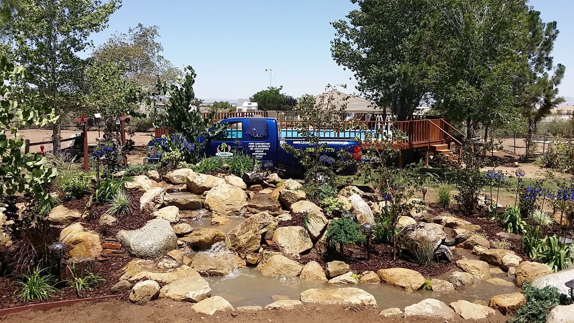 Rocky water feature in a garden with a blue truck parked in the background. Trees and plants surround the area.