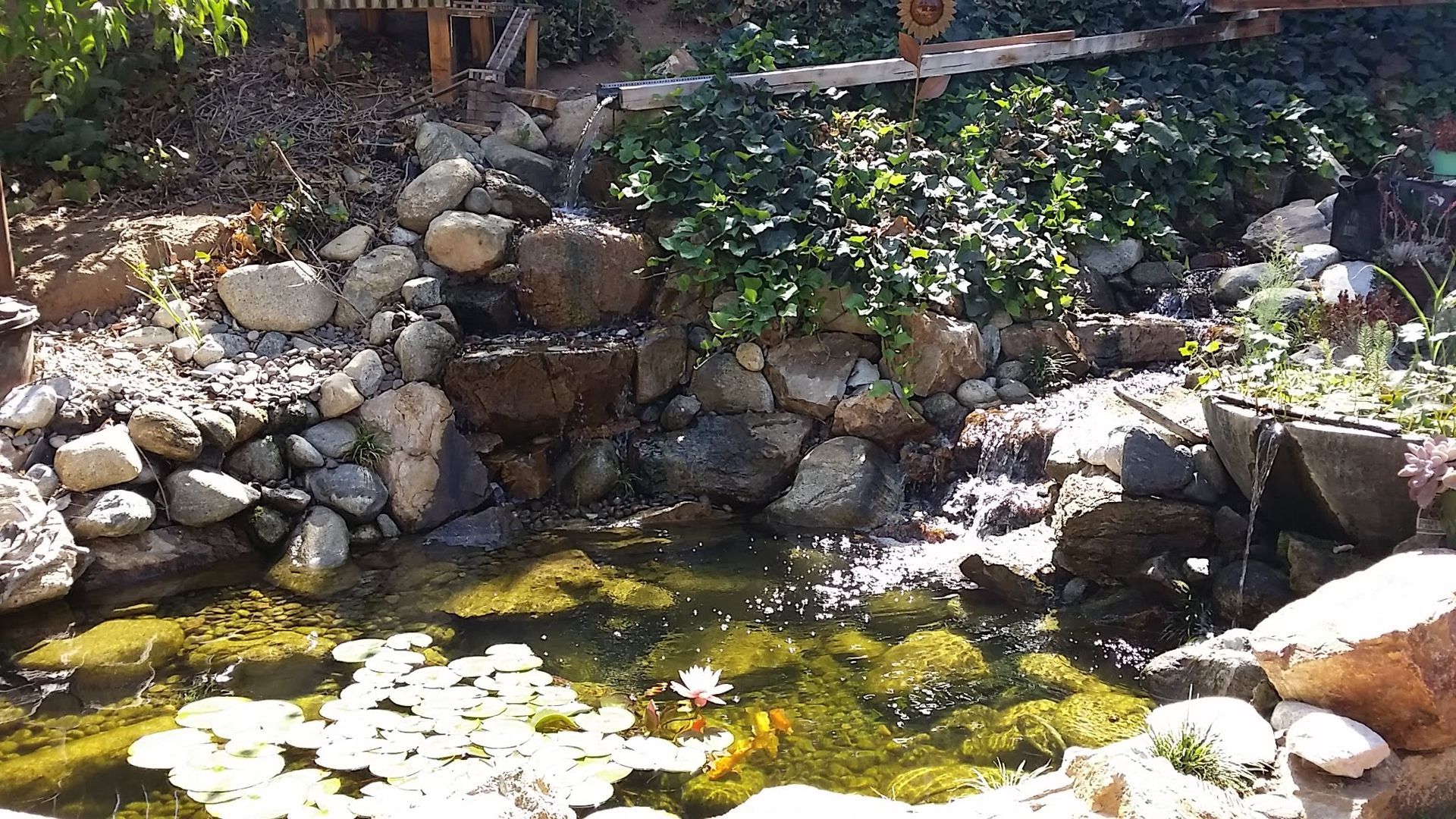 Stone waterfall cascading into a pond with lily pads and rocks, plants on top.