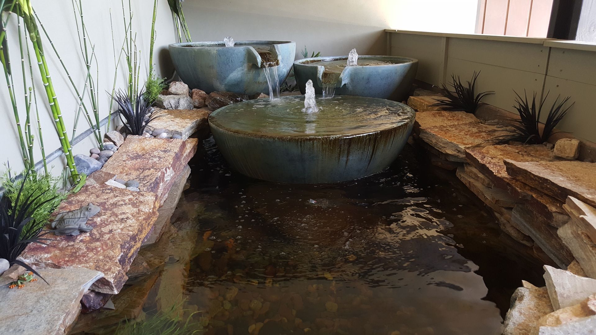 Water feature with three tiered, turquoise bowls and flowing water, surrounded by rocks and plants.