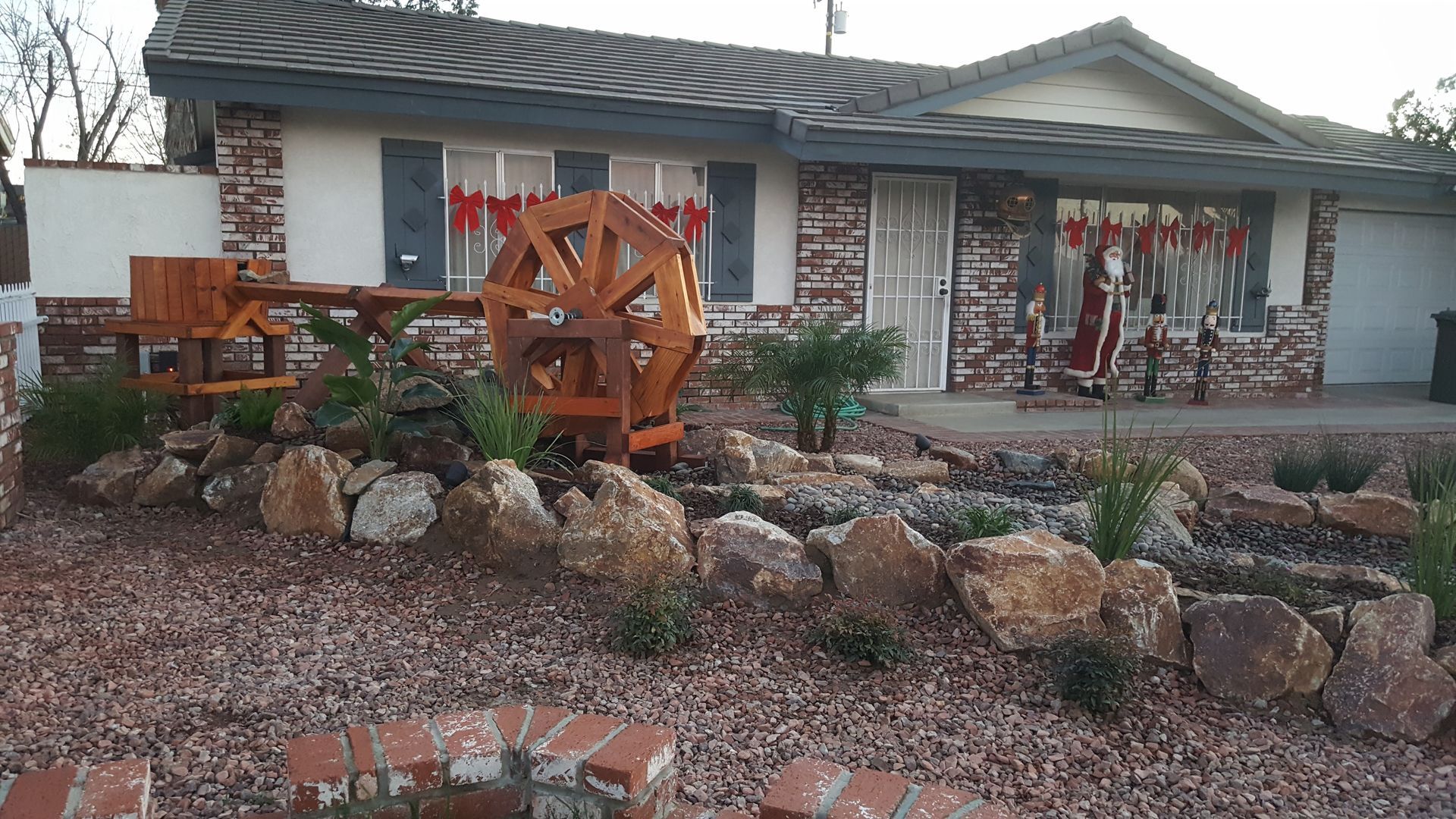 Small house with a water wheel feature in the front yard, surrounded by rocks and plants.