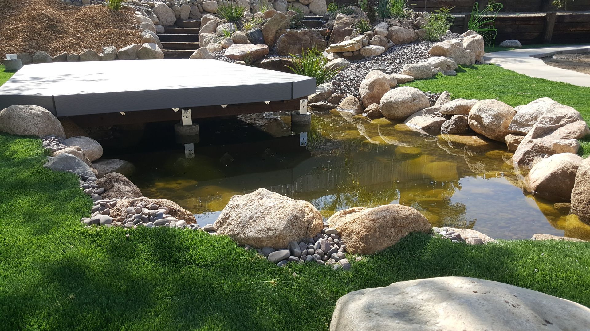 Pond with a gray deck, surrounded by rocks and green grass.