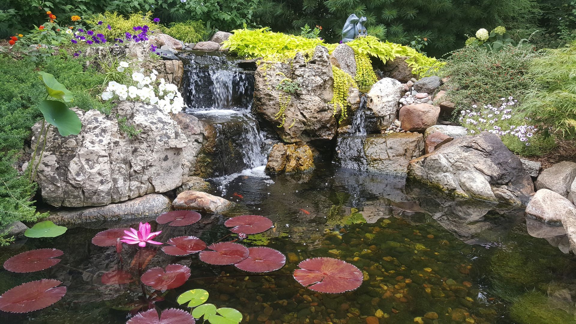 Small pond with waterfall, lily pads, and a pink water lily, surrounded by rocks and greenery.