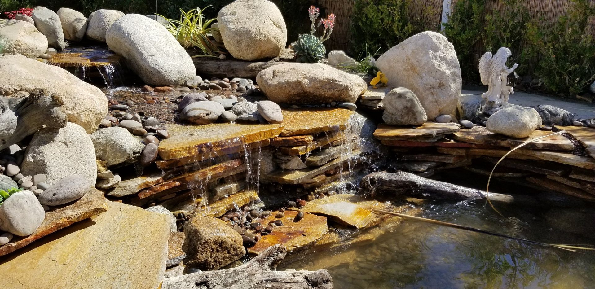 A small rock waterfall flows into a pond. Large stones and a statue of an angel are visible.