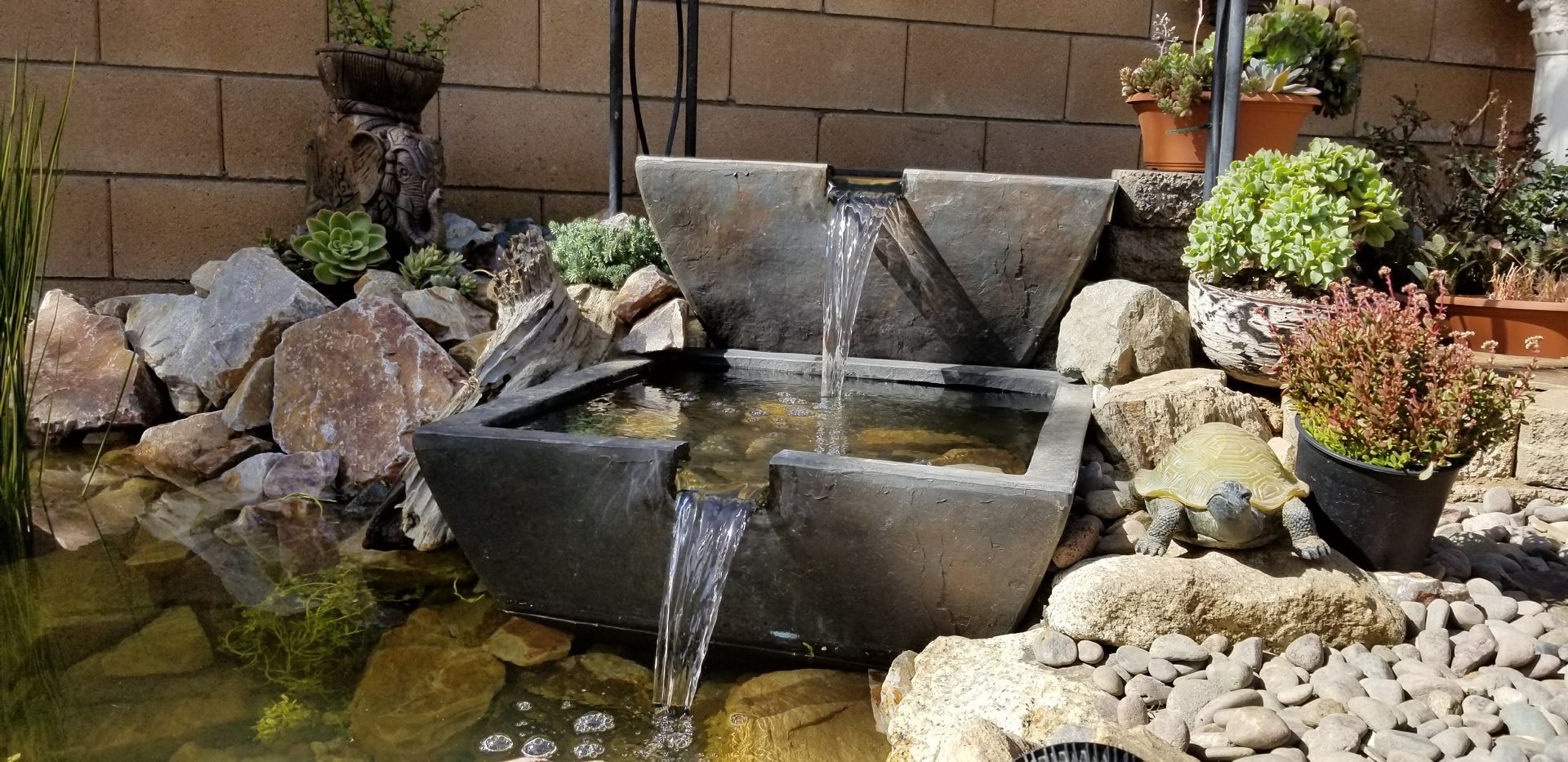 A tiered stone water fountain with flowing water, surrounded by rocks and plants.