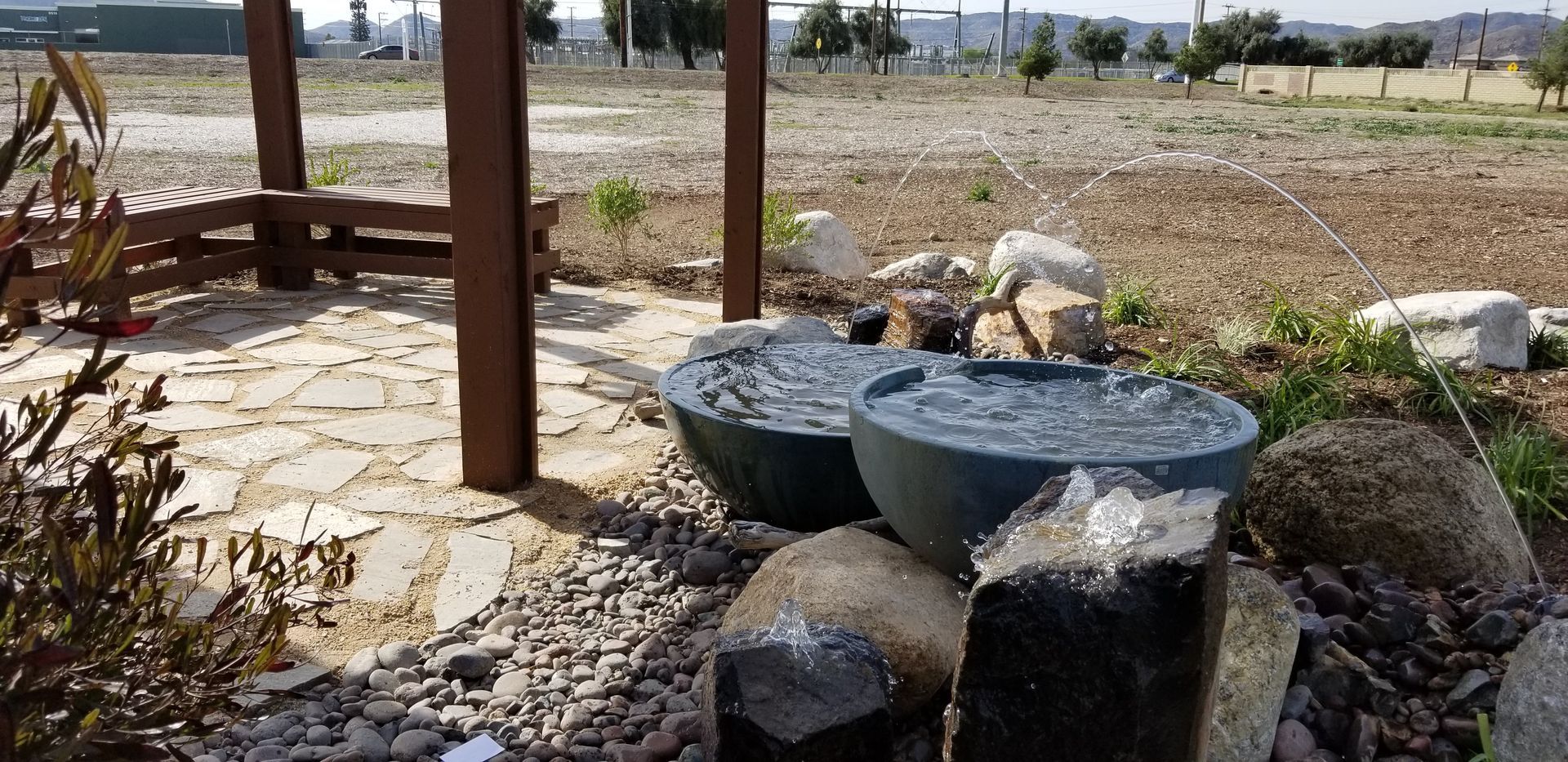 Water fountain with two dark bowls, flowing water, and large rocks in a garden setting.