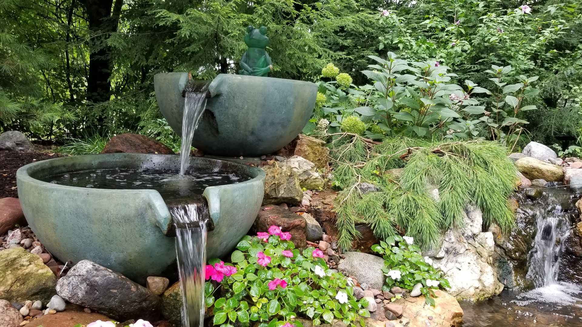 Water fountain in a garden with tiered bowls, overflowing into a small pond. Greenery and flowers surround.