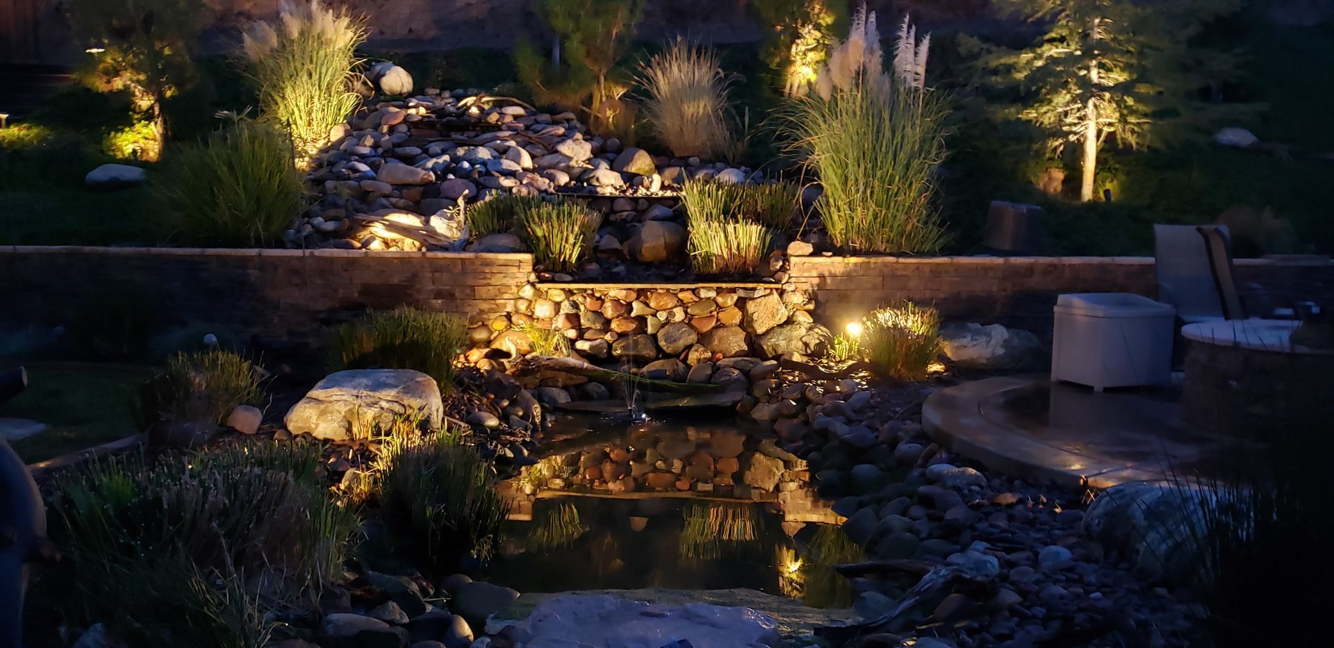 A night shot of a decorative garden with a water feature, illuminated by warm lights.
