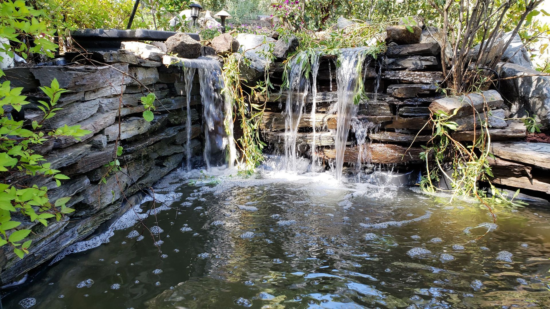 Small waterfall cascading into a pond, built of layered stone, with greenery.