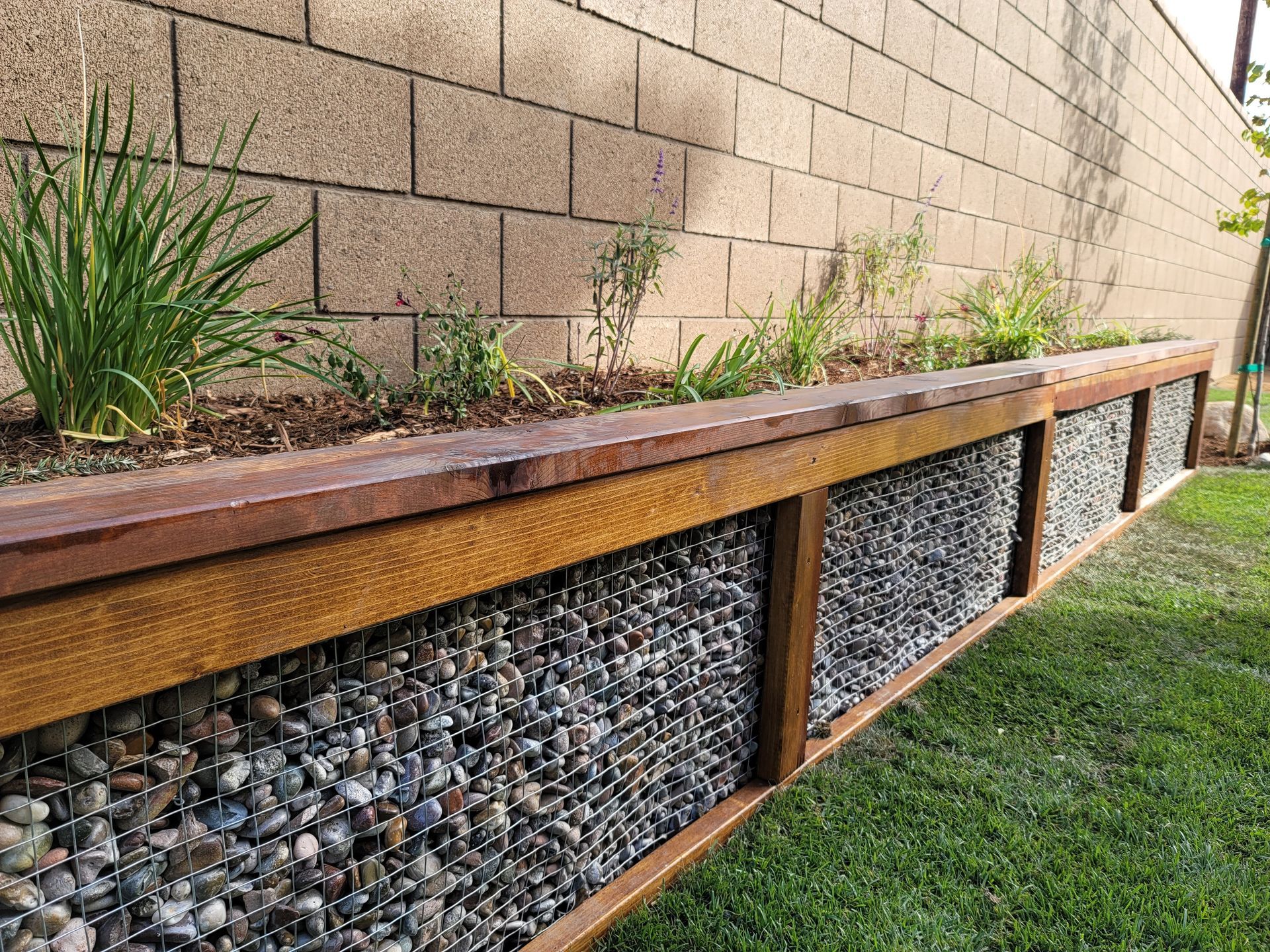 Raised garden bed with wood frame and wire mesh filled with rocks, against a brick wall. Green plants on top.