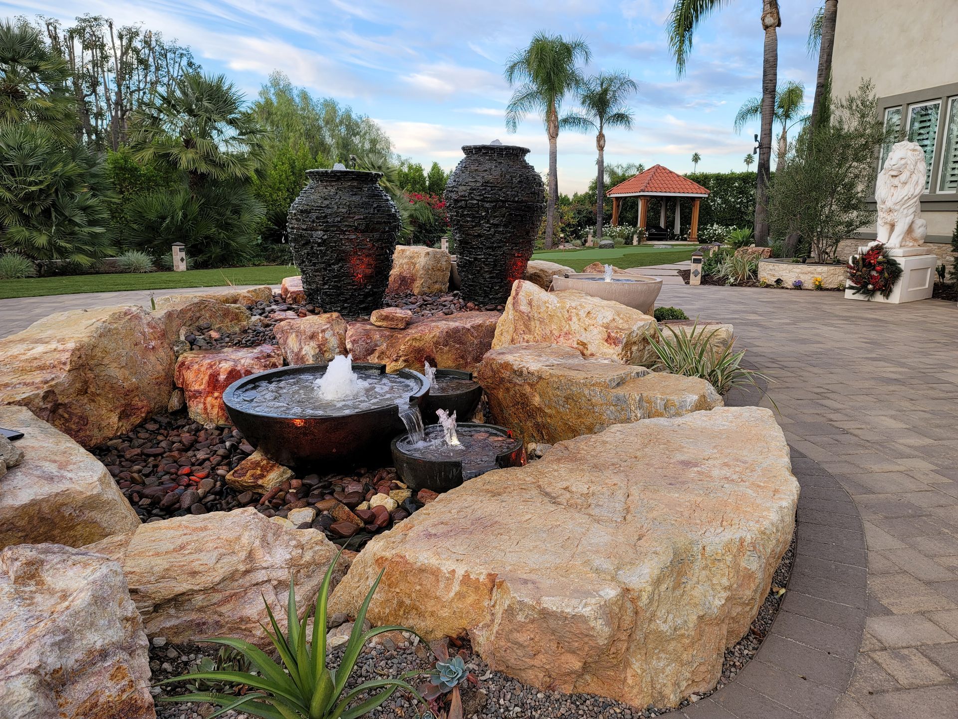 Water fountain with three dark urns, a bowl with water jets, surrounded by rocks and a garden.