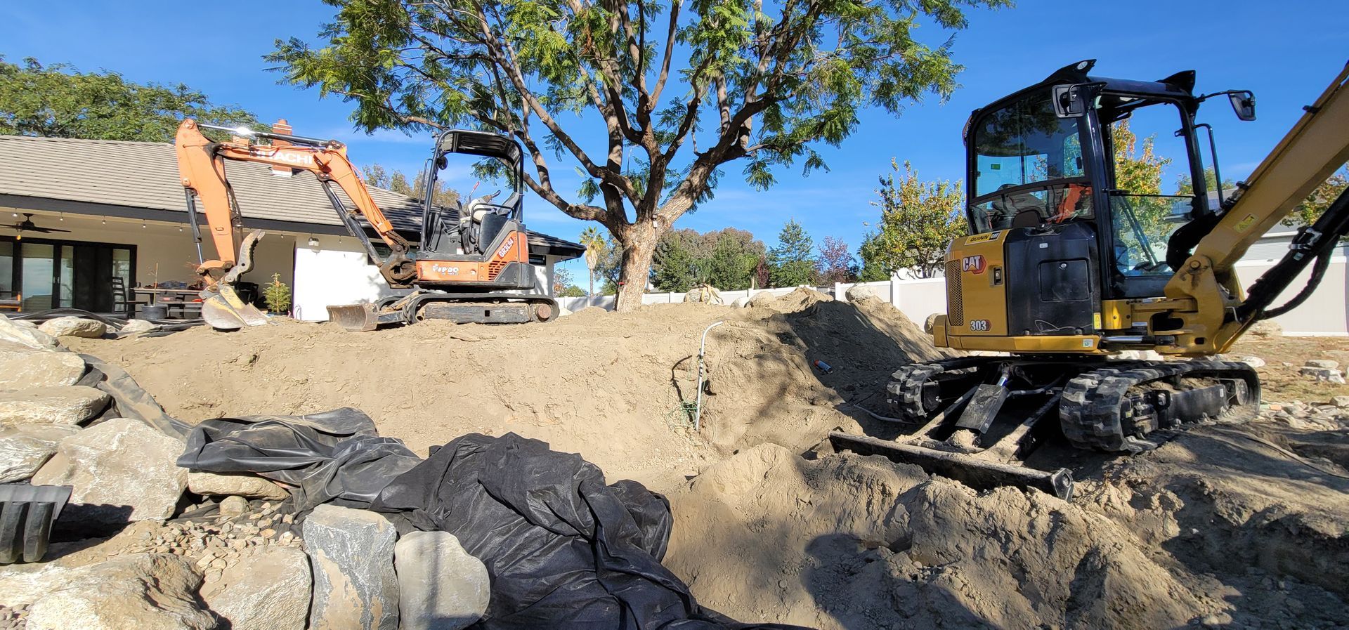 Two excavators at a construction site, moving dirt and debris next to a house under a clear sky.