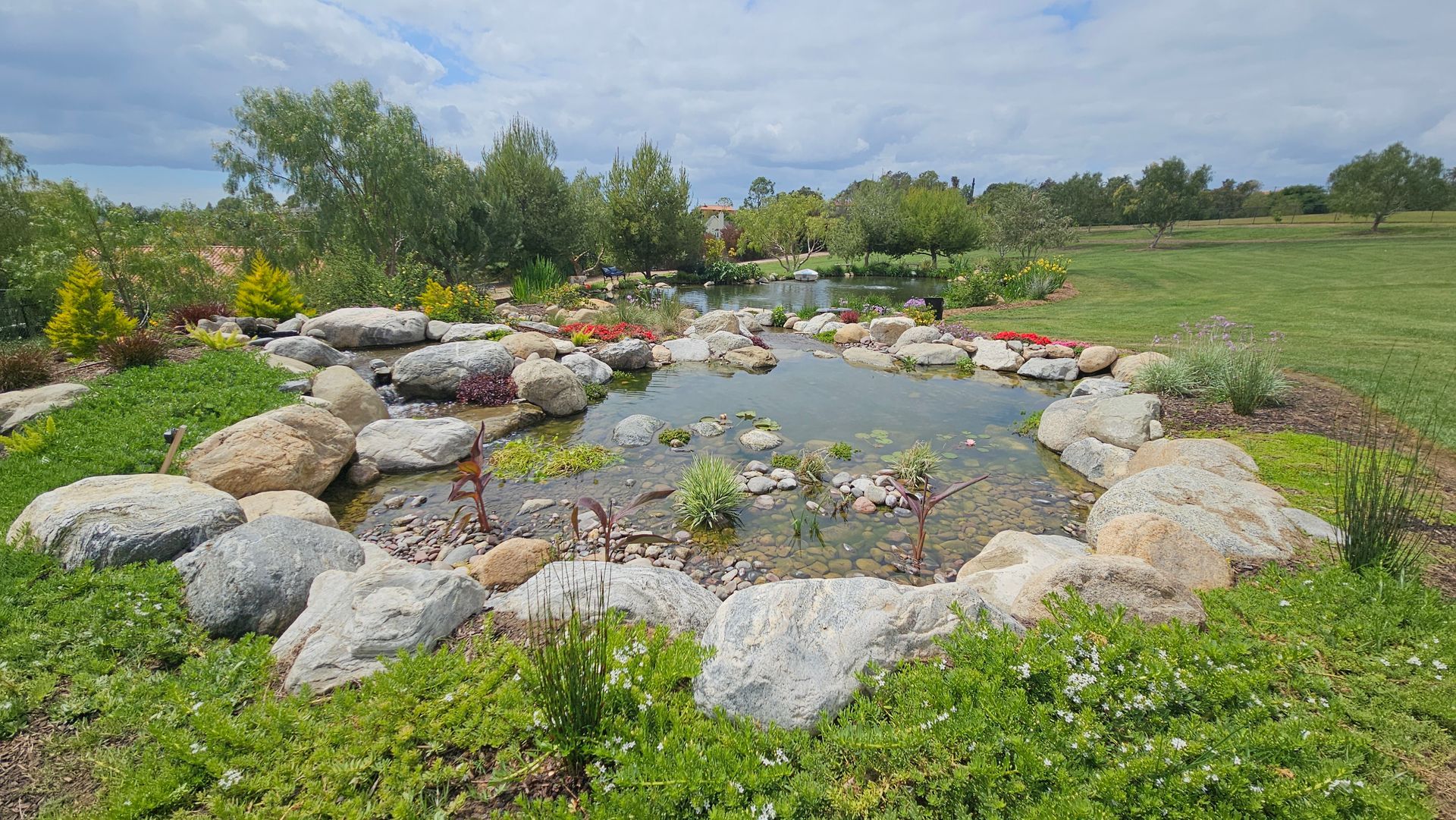 Small pond surrounded by rocks and greenery in a grassy landscape under a cloudy sky.