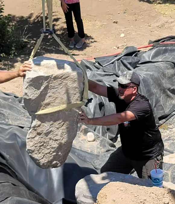 Man and assistant positioning a large rock into a pond with lifting straps.