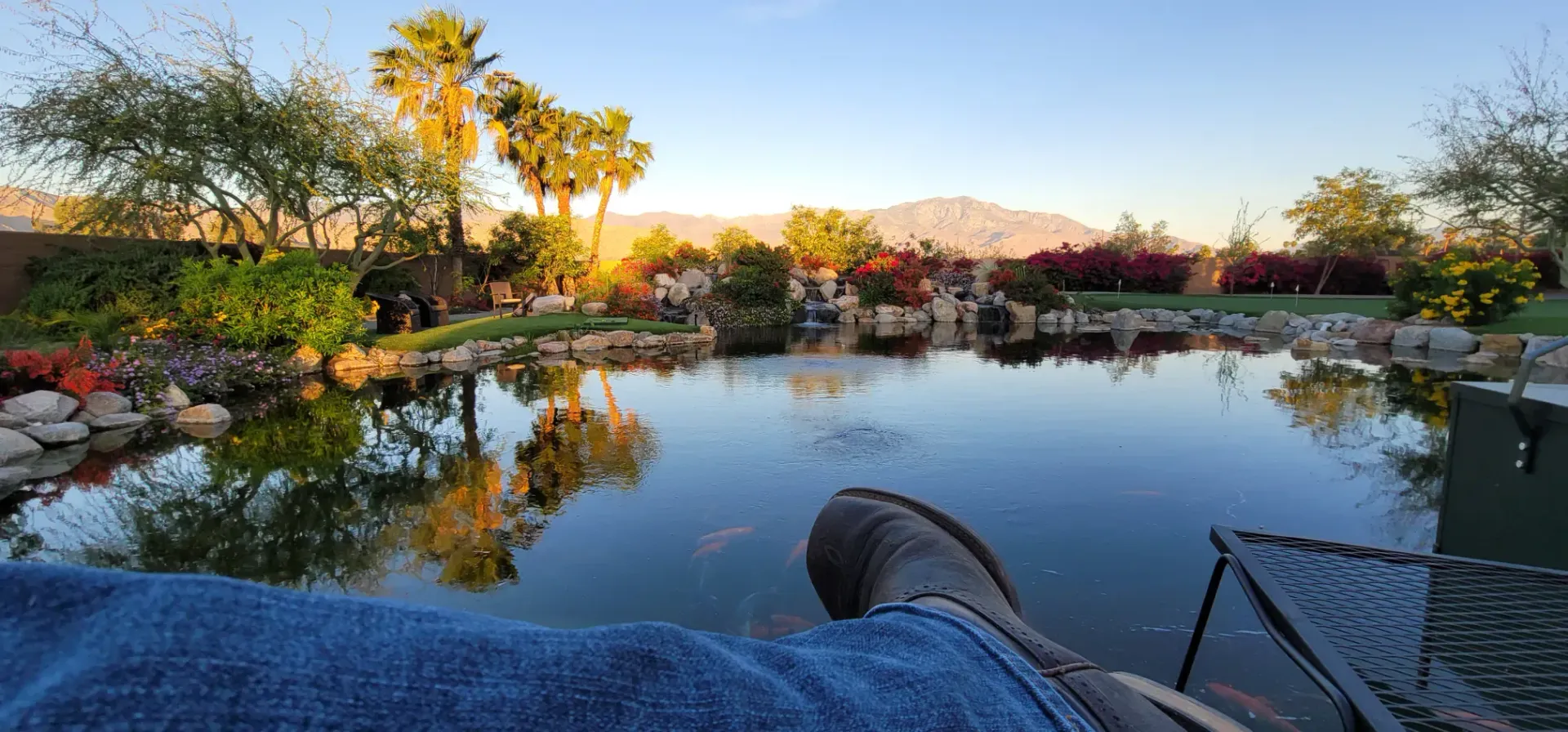 Person relaxing by a pond, with mountains and trees in the background. Calm water reflects the sky and trees.