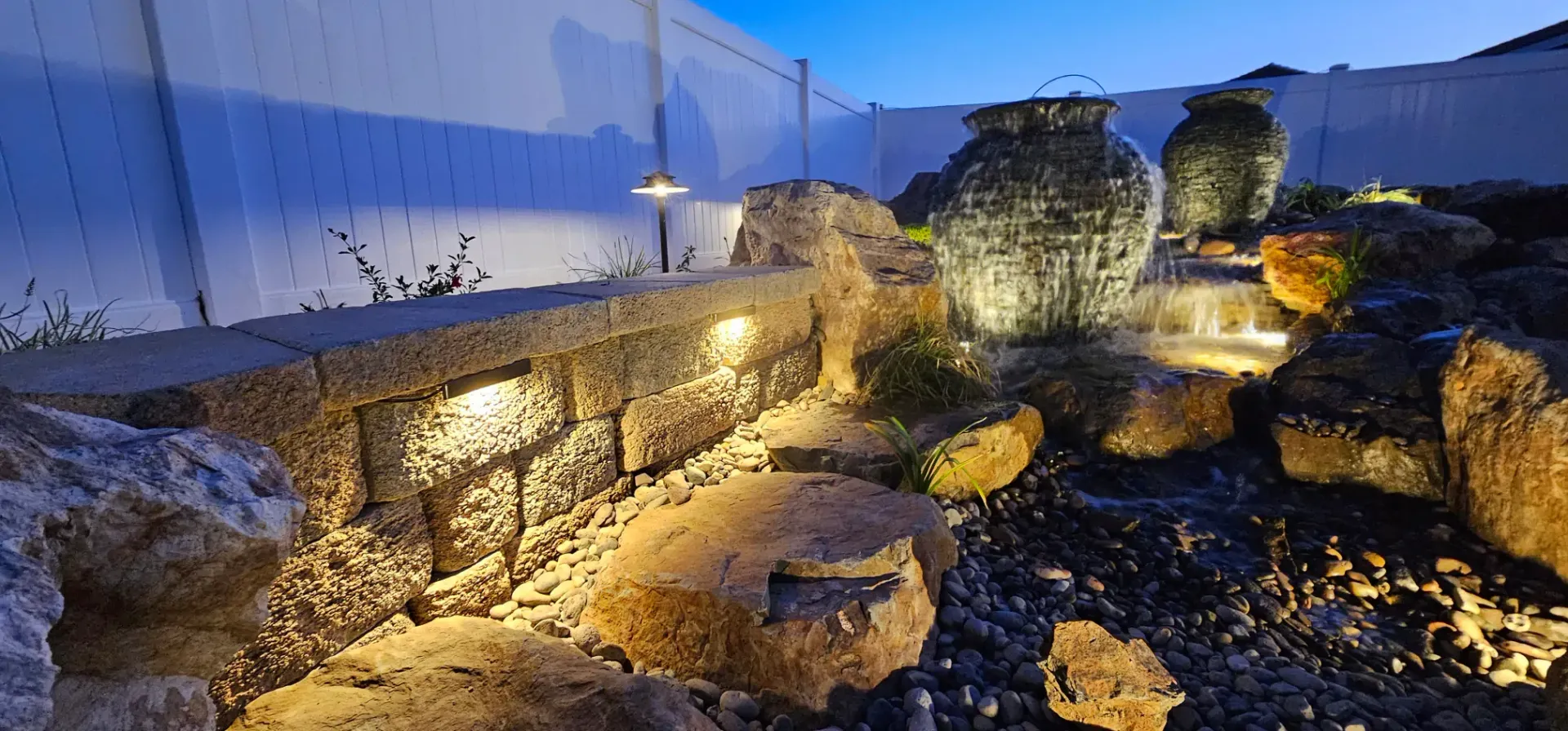 Stone water feature with glowing lights in a backyard. A decorative wall is in the foreground.