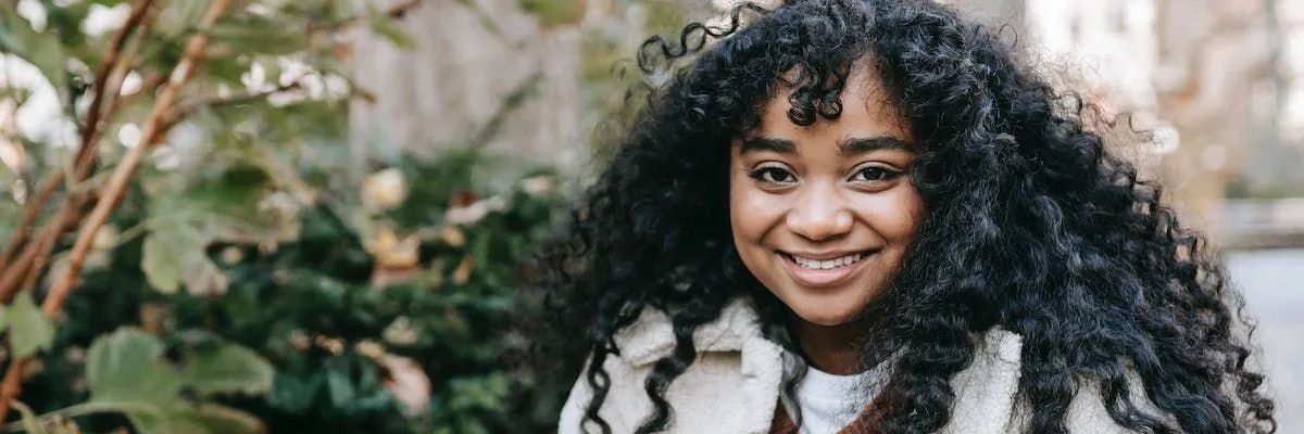 A woman with curly hair is smiling in front of a tree.