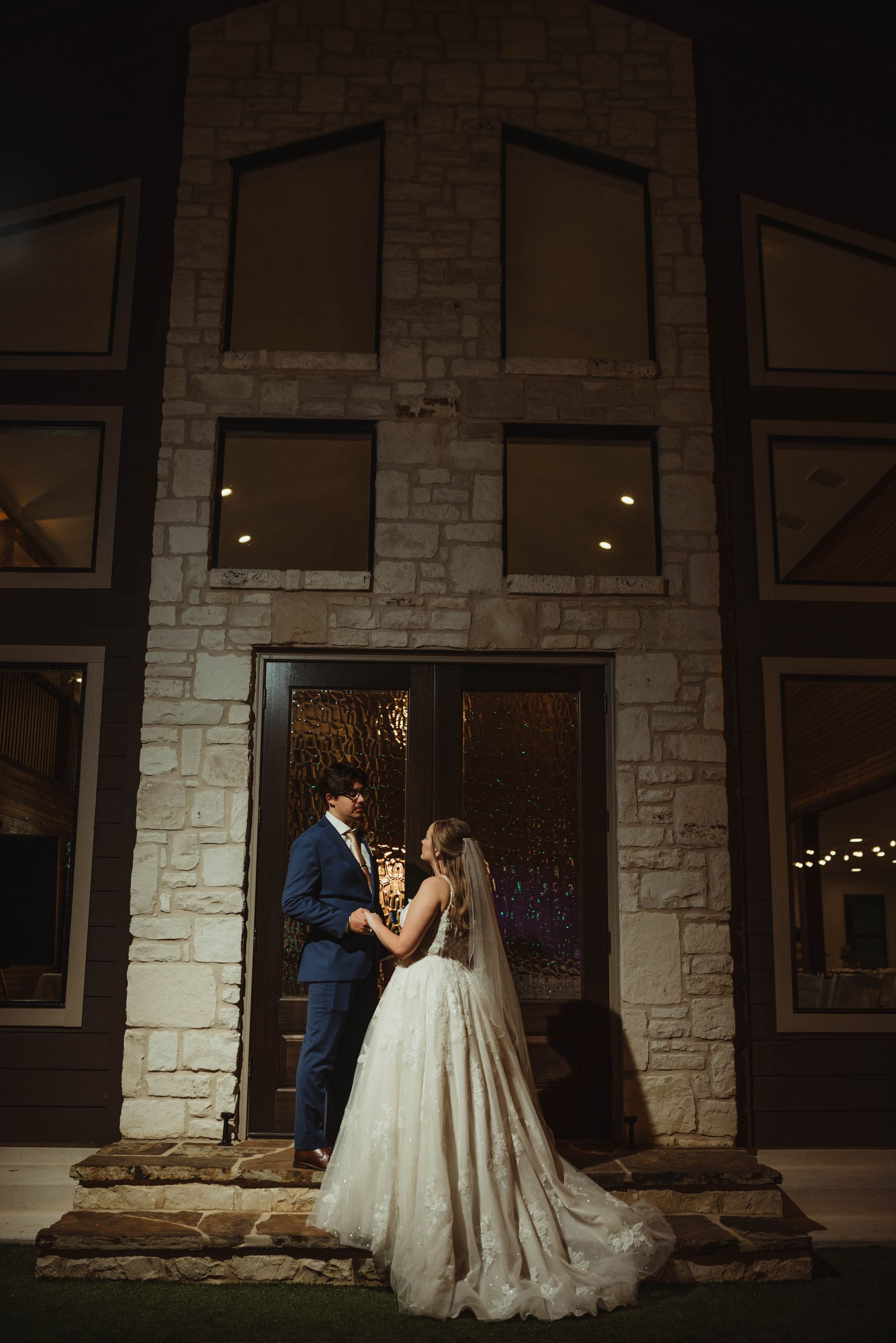 Couple in wedding attire stand before a stone building with glass doors, illuminated at night.