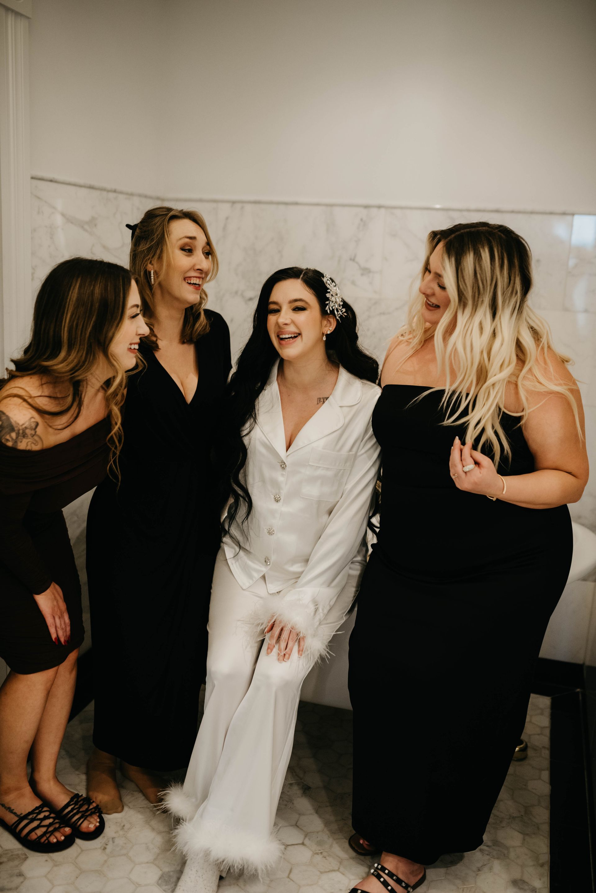 Bride in white pajamas laughs with bridesmaids in black dresses in a bathroom.