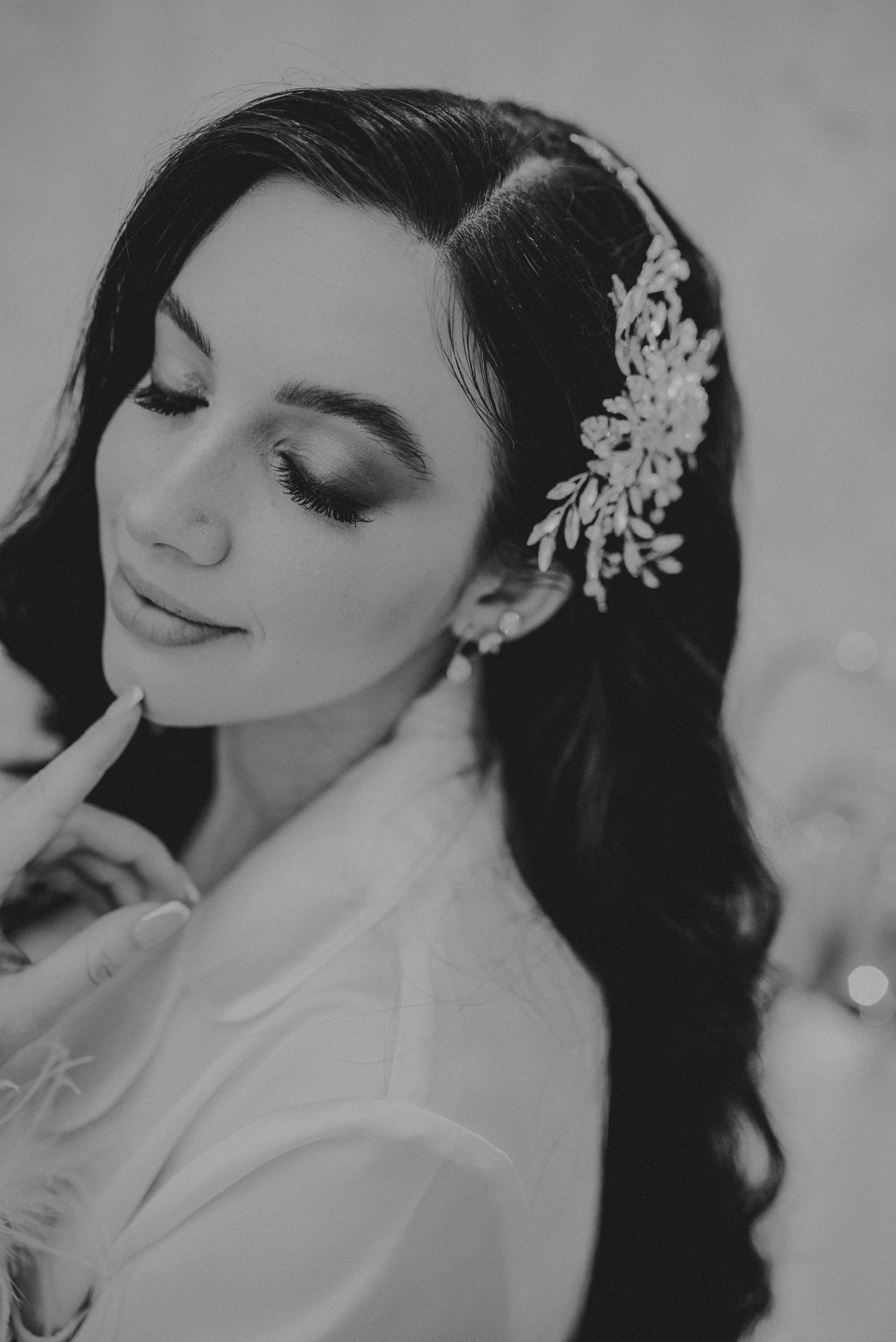 Woman with dark hair and embellished hairpiece, wearing a robe, looking down, touching her chin.