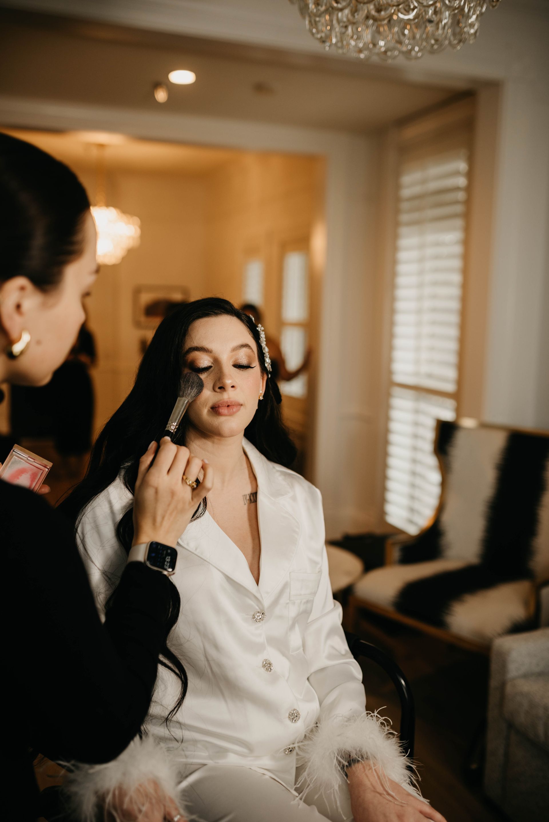 A woman has makeup applied while seated in a well-lit room. She wears white pajamas with feather trim.