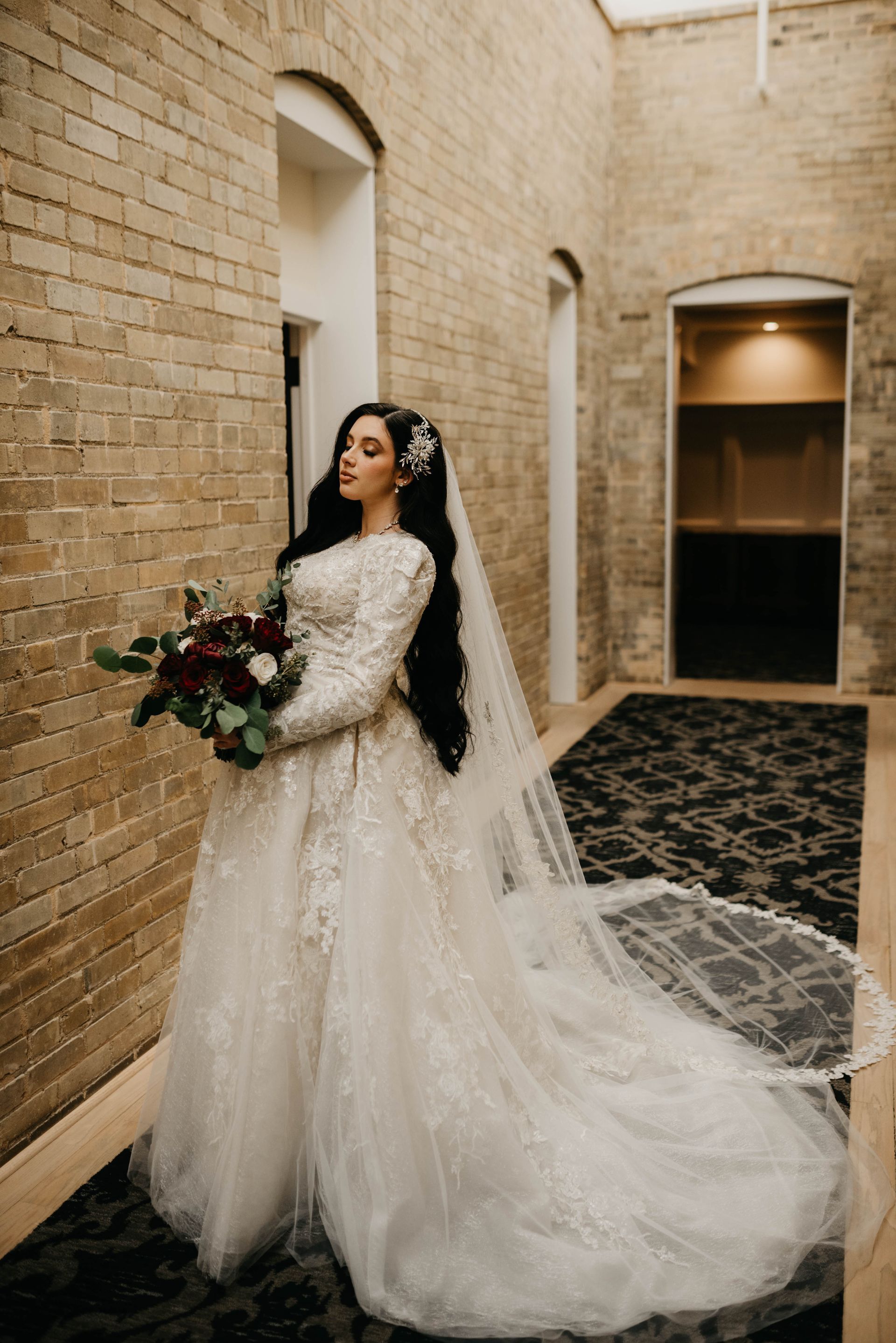 Bride in a long-sleeved, embroidered gown with a long veil, holding a bouquet. Standing in a hallway.