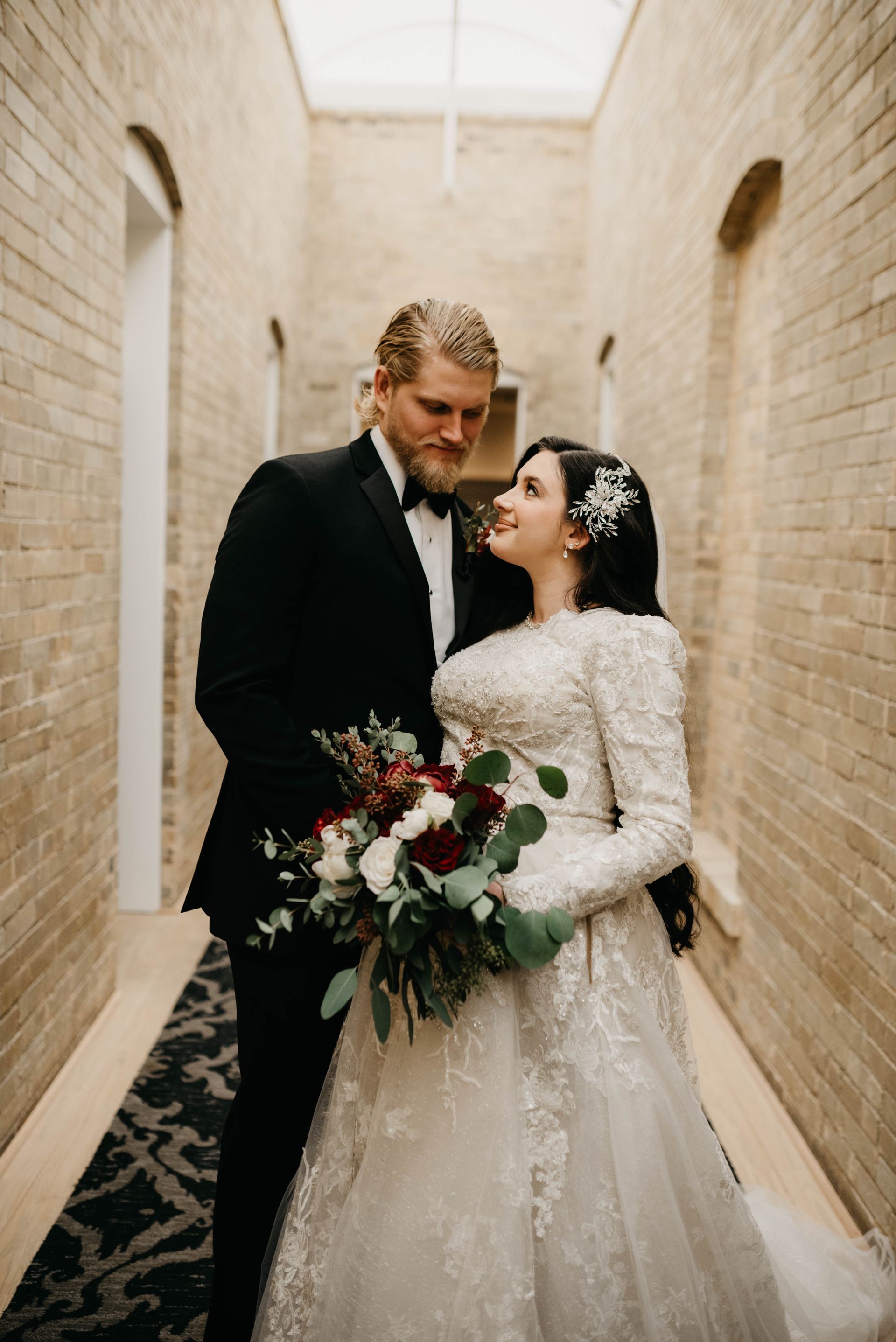 Bride and groom embrace in a brick hallway, holding flowers. The bride wears a long-sleeved dress, and the groom is in a tuxedo.