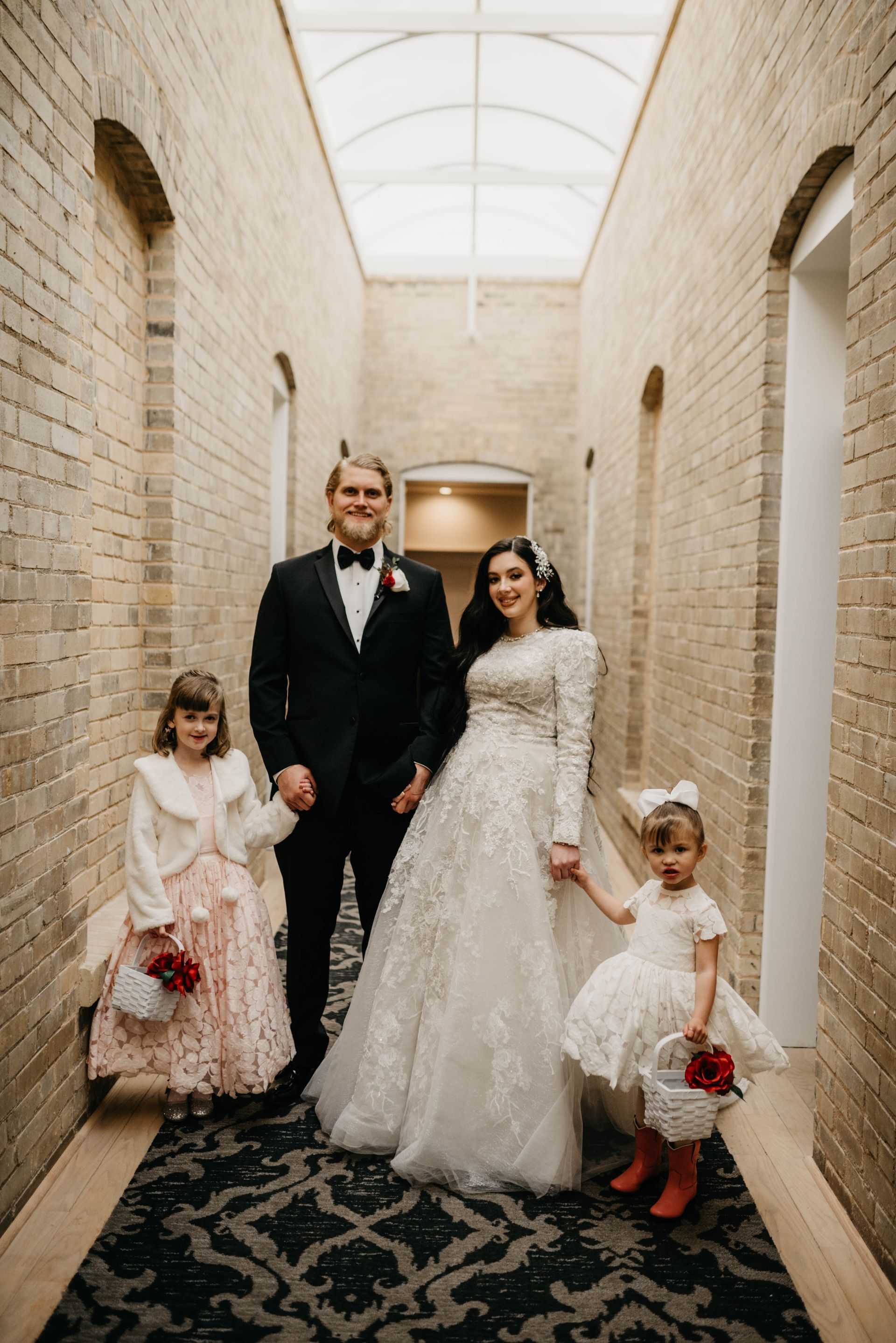 Bride, groom, and flower girls pose in a brick hallway. The bride wears a white gown. The groom wears a black tuxedo.