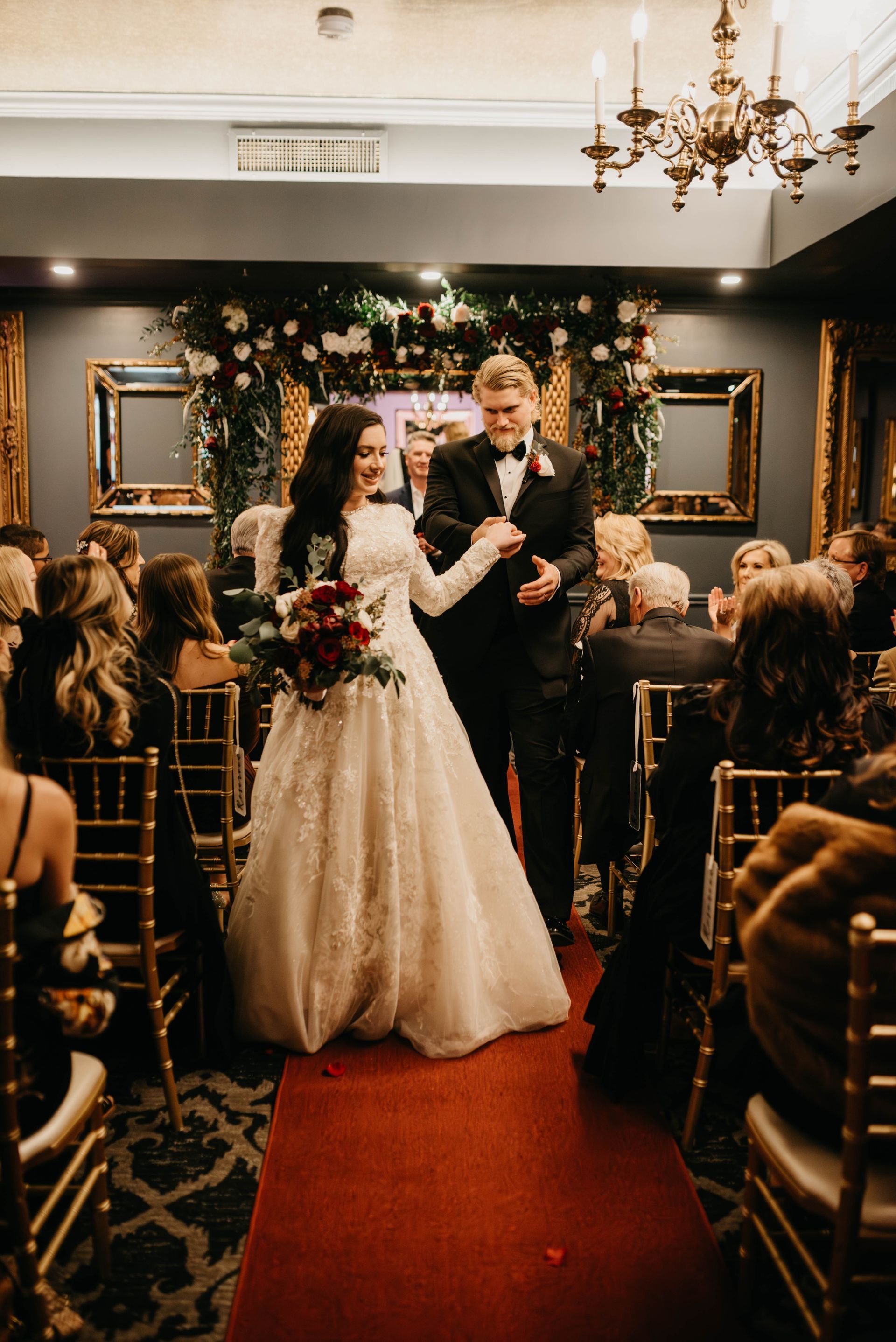 Bride and groom walk down the aisle after wedding ceremony, red carpet, floral arch, gold chairs.