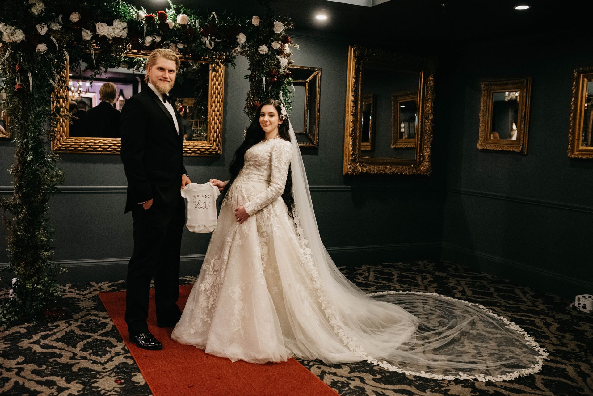 Groom and pregnant bride pose at wedding; archway flowers and gold-framed mirrors in the background.